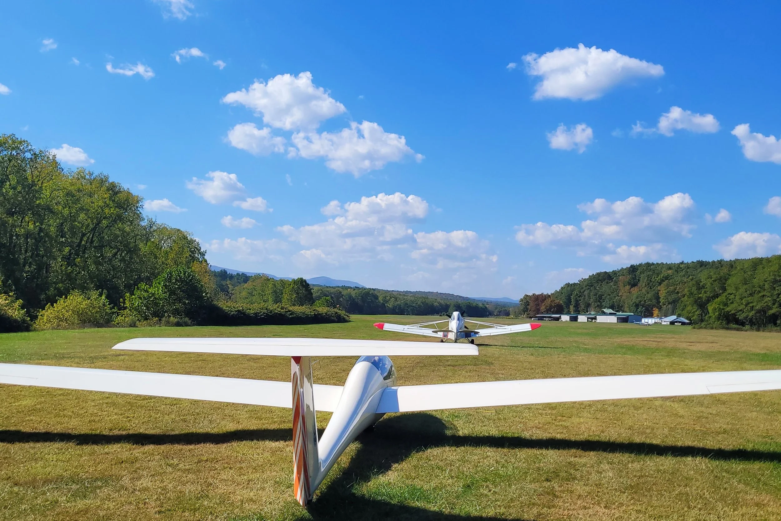 A glider lined up behind a tow plane ready for take-off on a grassy field with trees and hangars in the background under a blue sky with clouds.