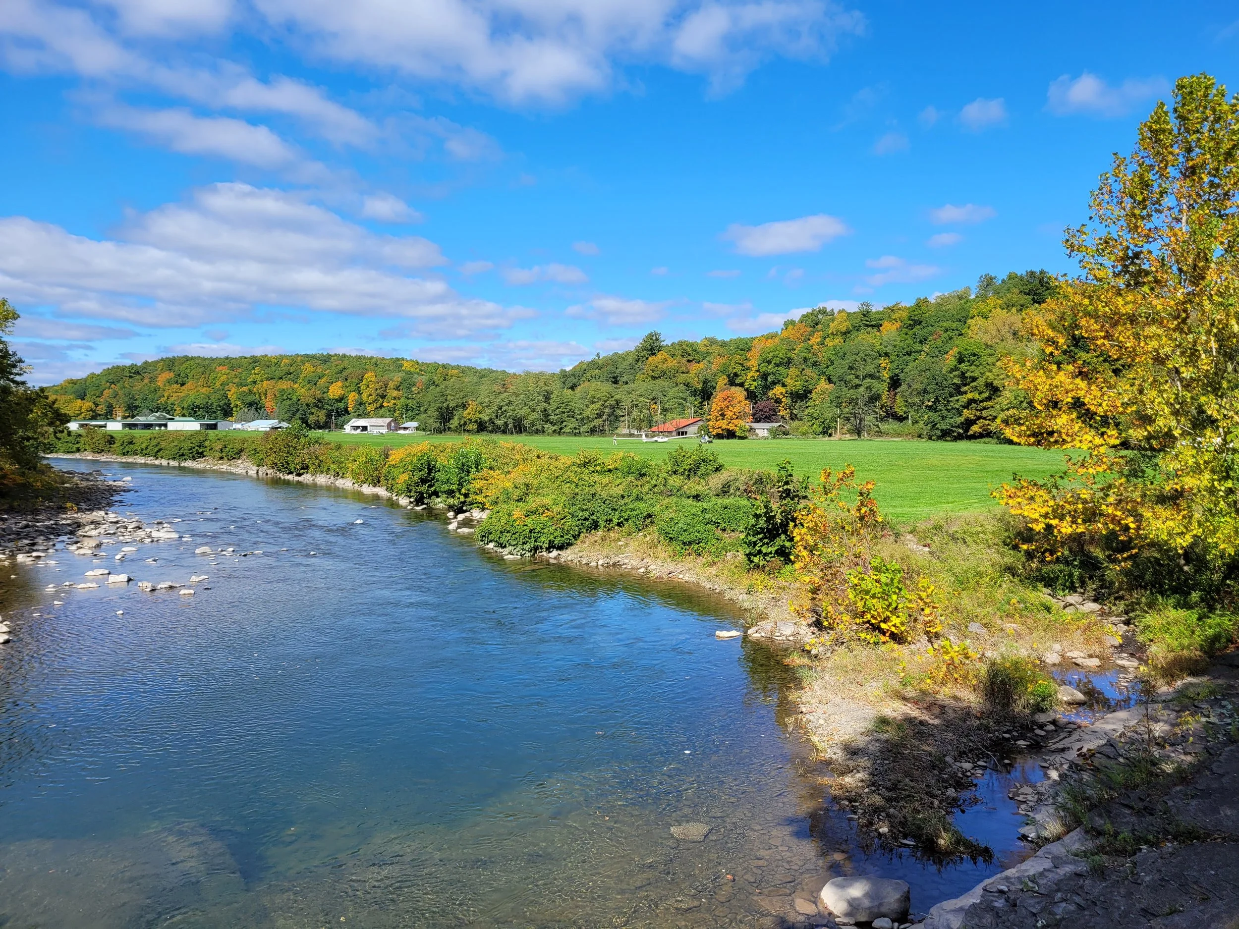 A sunny Freehold Airport landscape with the Catskill creek flowing along the green grass airstrip and trees showing fall colors, with a blue sky and white clouds overhead.