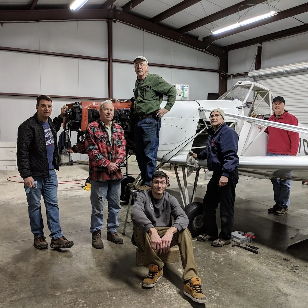Six men standing and sitting around our Pawnee tow plane inside a hangar, working on the plane's engine.