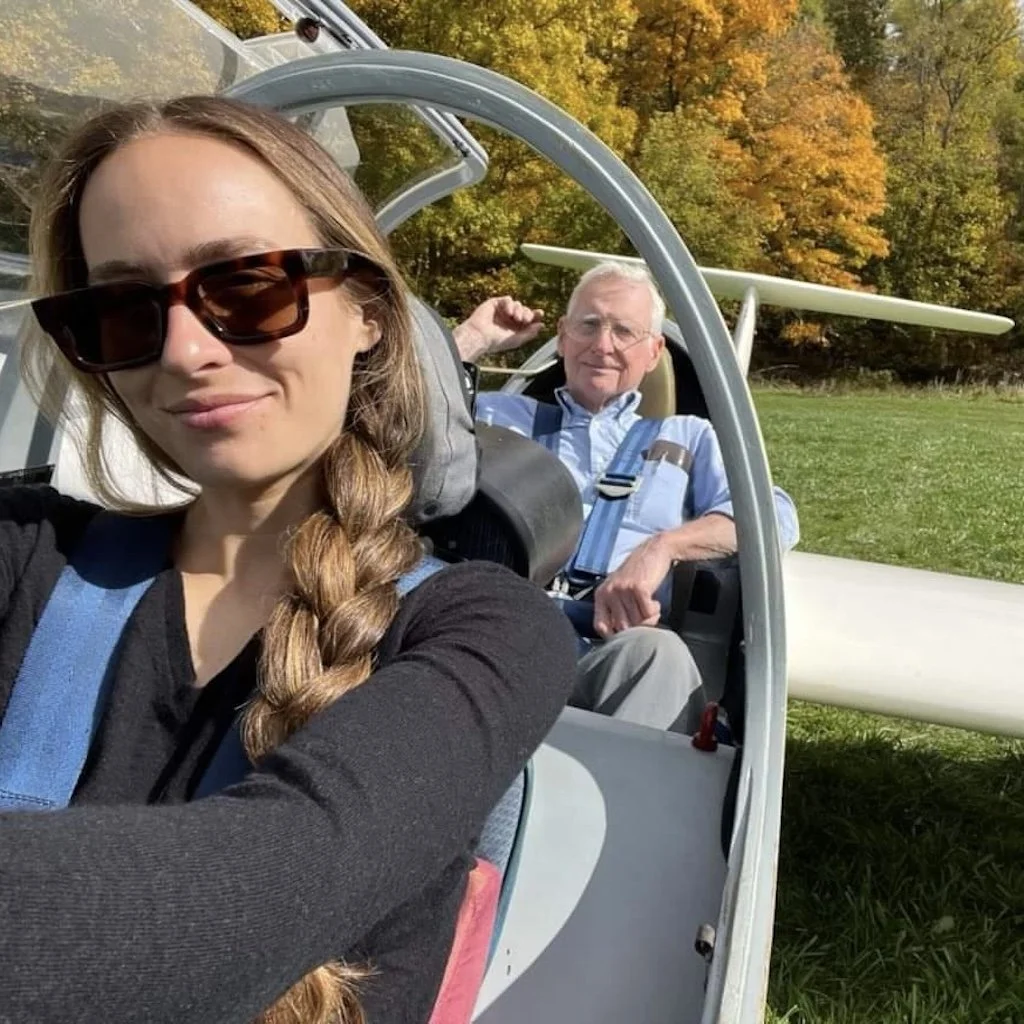 Tailor and Randol sitting inside a glider waiting for take-off, with a background of autumn trees and green grass.