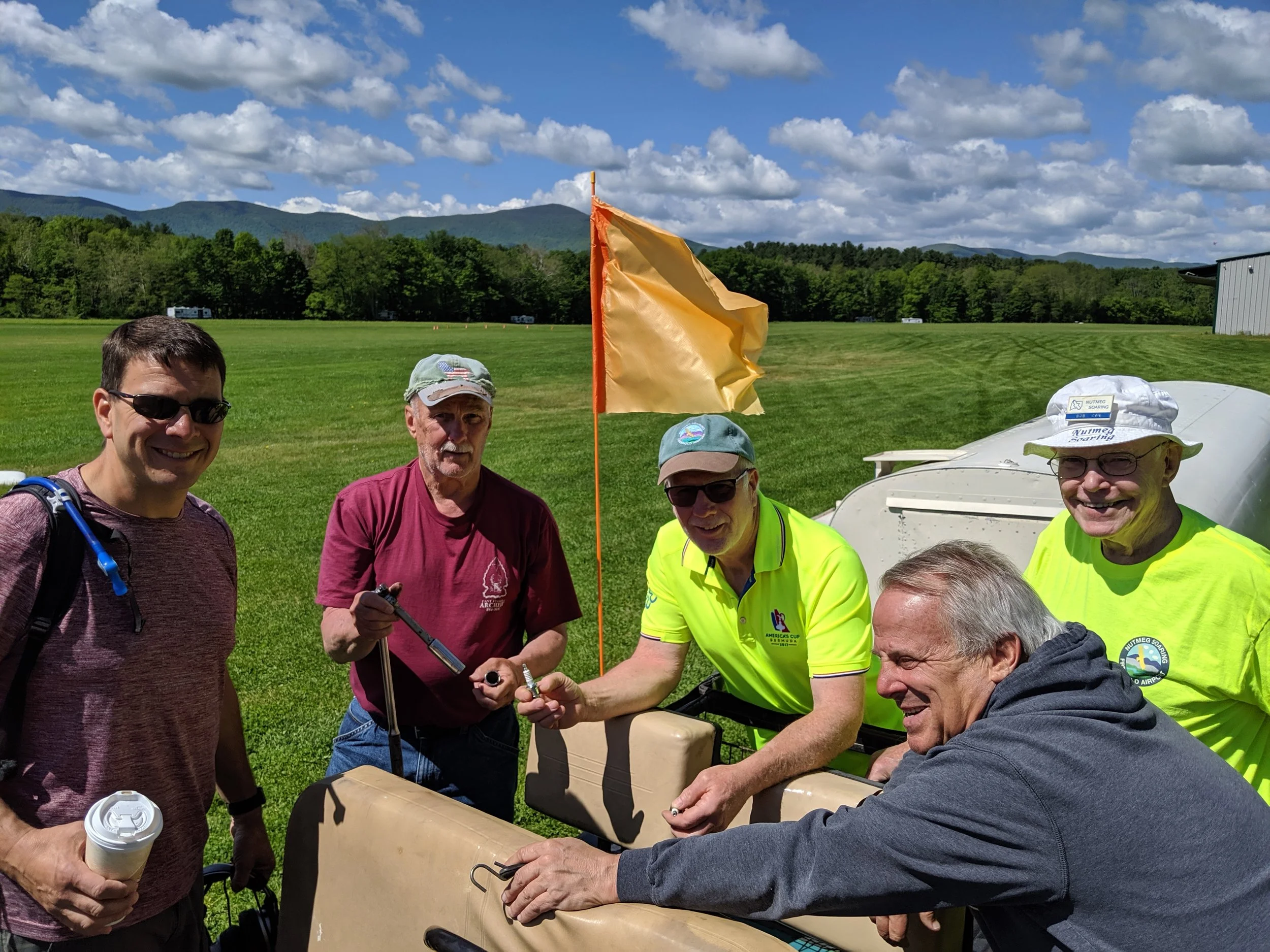 Group of Nutmeg members on a grassy field, some wearing sunglasses and bright yellow shirts, gathered around a golf cart, with a yellow flag on a pole nearby, and mountains in the background.