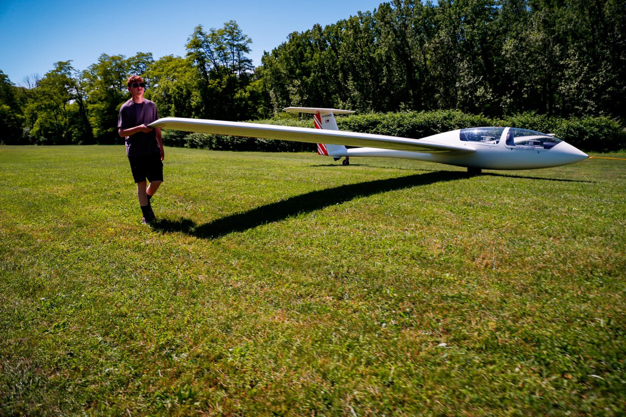 A young man walks along a glider holding its wing at it is towed on a grassy field on a sunny day.