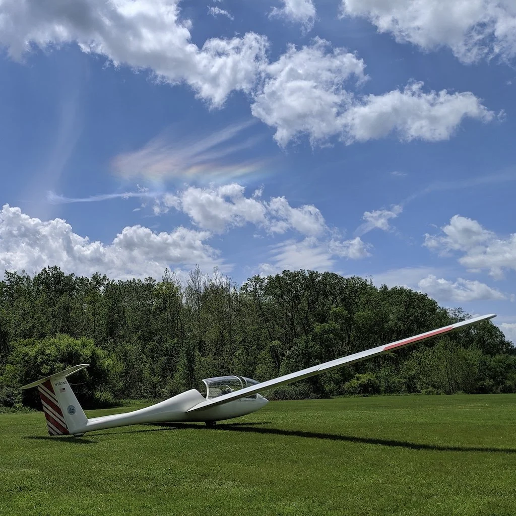 A sailplane resting on a grassy field with a backdrop of trees and a blue sky with clouds and a rainbow-colored optical phenomenon.