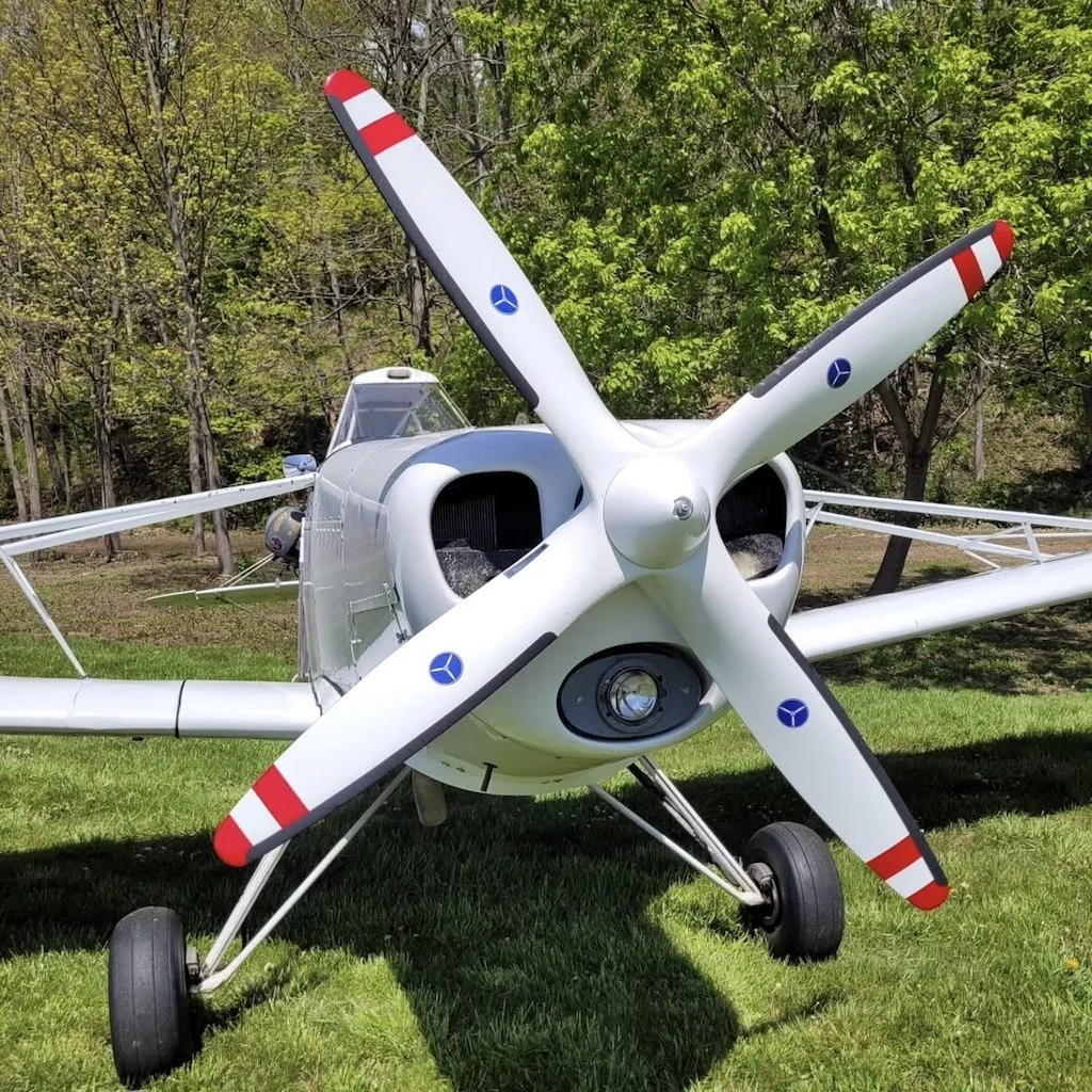 Close-up front view of a Pawnee tow plan with a white body and a four-blade propeller marked with red tips and blue logos, parked on grass with trees in the background.