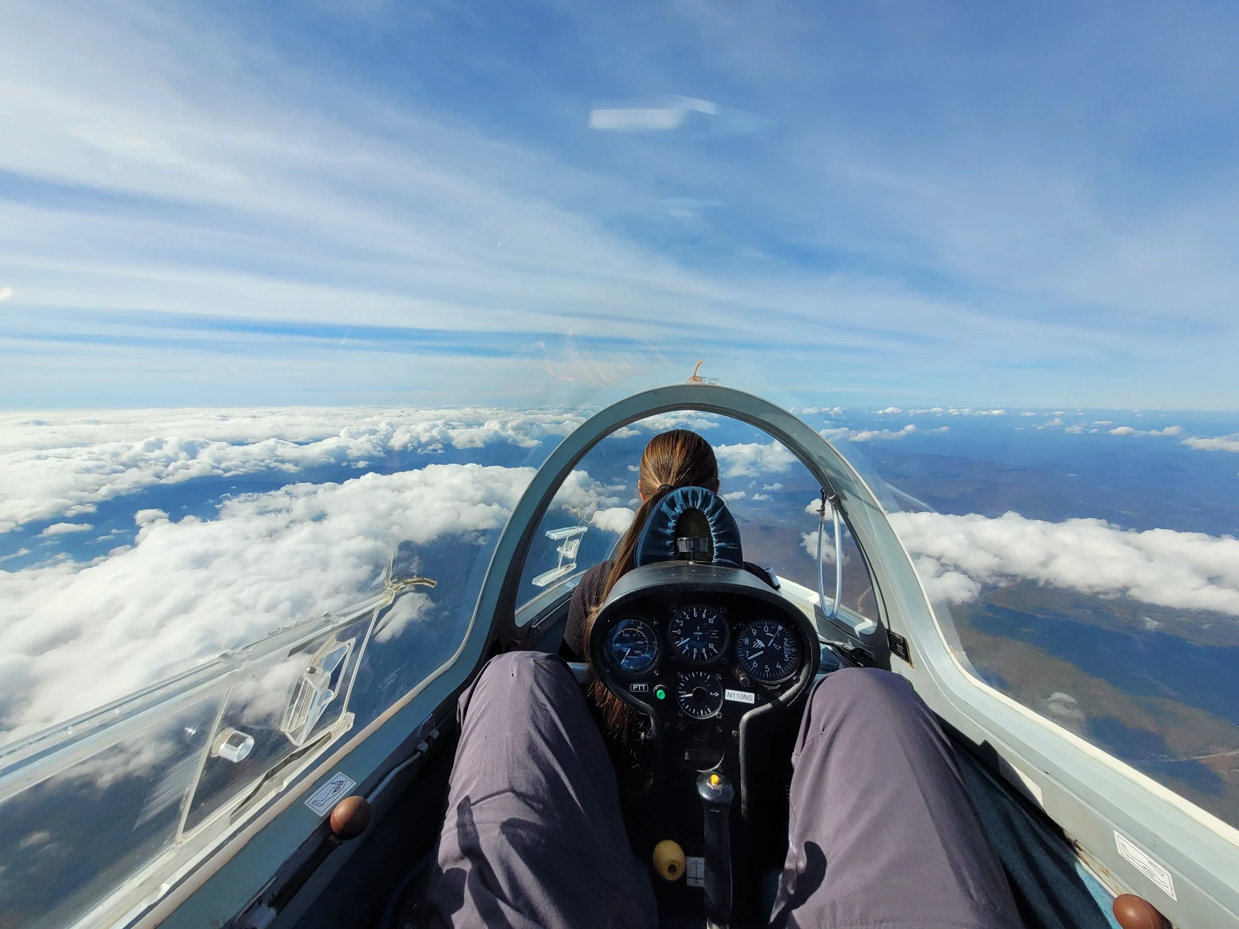 View from inside a sailplane cockpit looking out at blue sky and clouds, with the pilot's head  visible.
