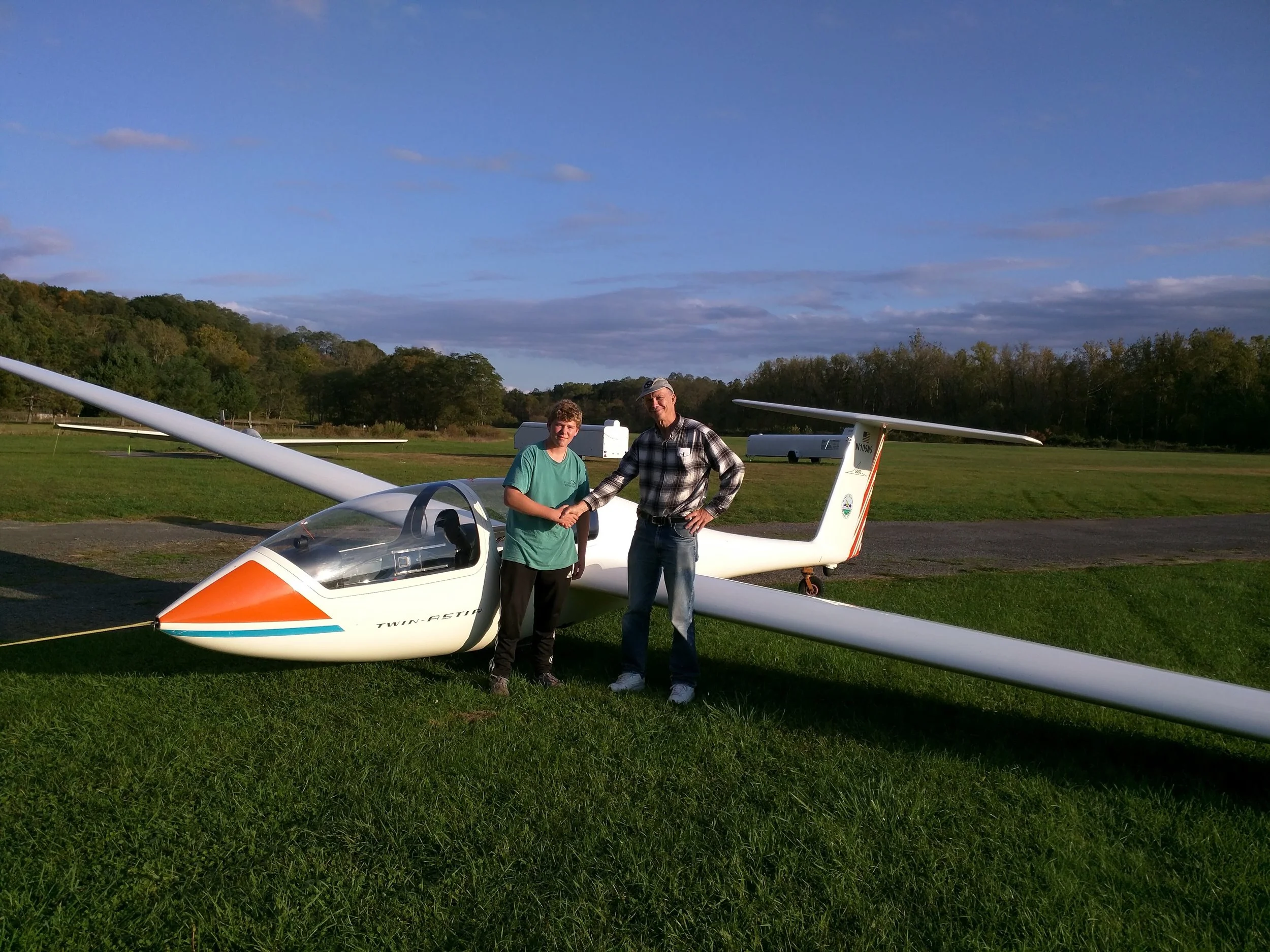 Chris and Marty shake hands in front of a white glider on a grassy field, with a clear blue sky and trees in the background. Chris just completed his first Solo.