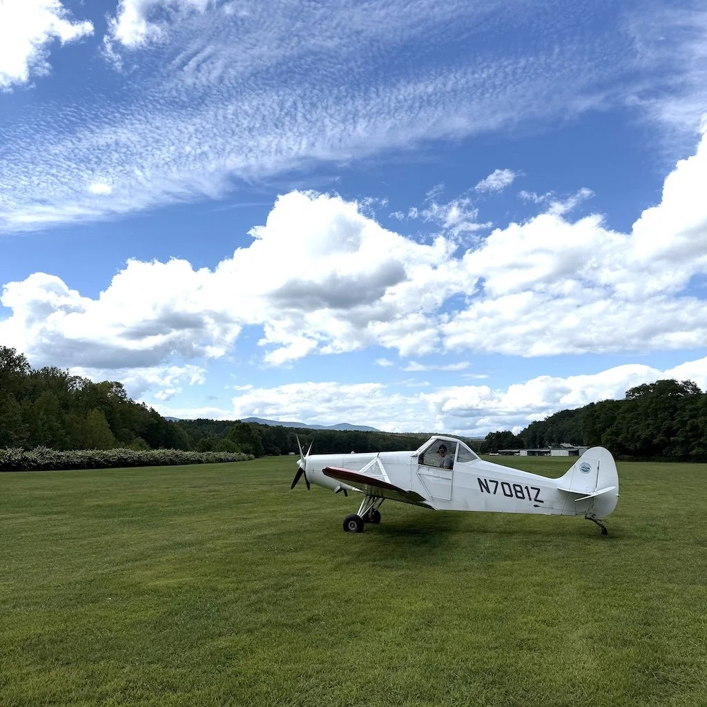 A Pawnee tow plane parked on a grassy field with a blue sky and white clouds overhead.