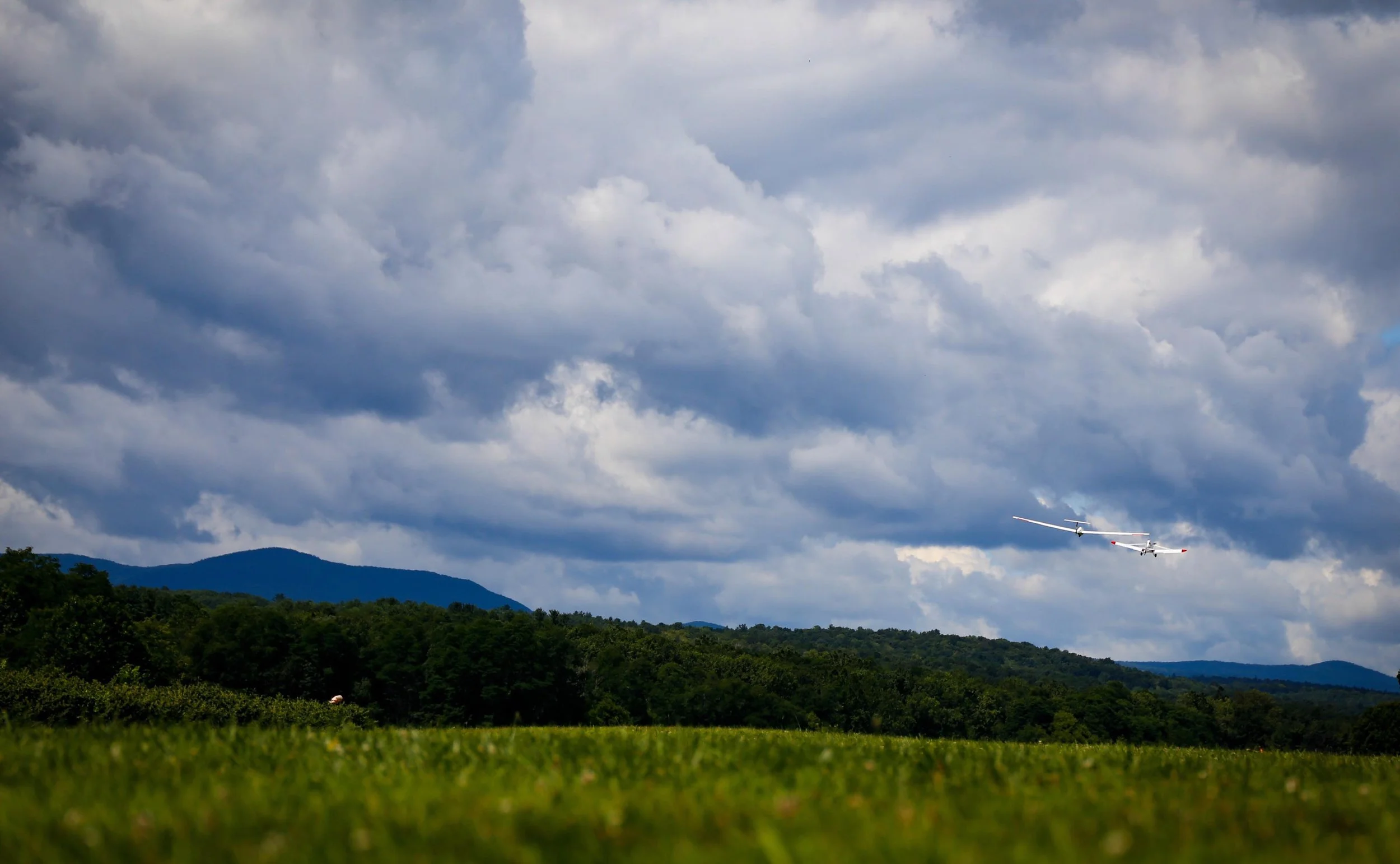 A gliders being towed by an airplane flying over a grassy field with wooded hills and cloudy sky in the background.