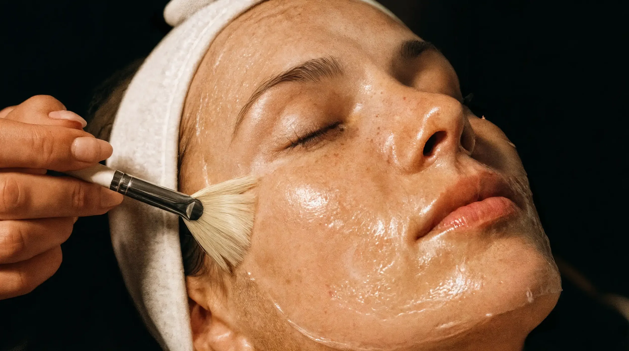Una donna riceve un trattamento di cura del viso, con un professionista che applica un prodotto con un pennello sulla sua pelle liscia e idratata, mentre è distesa con gli occhi chiusi