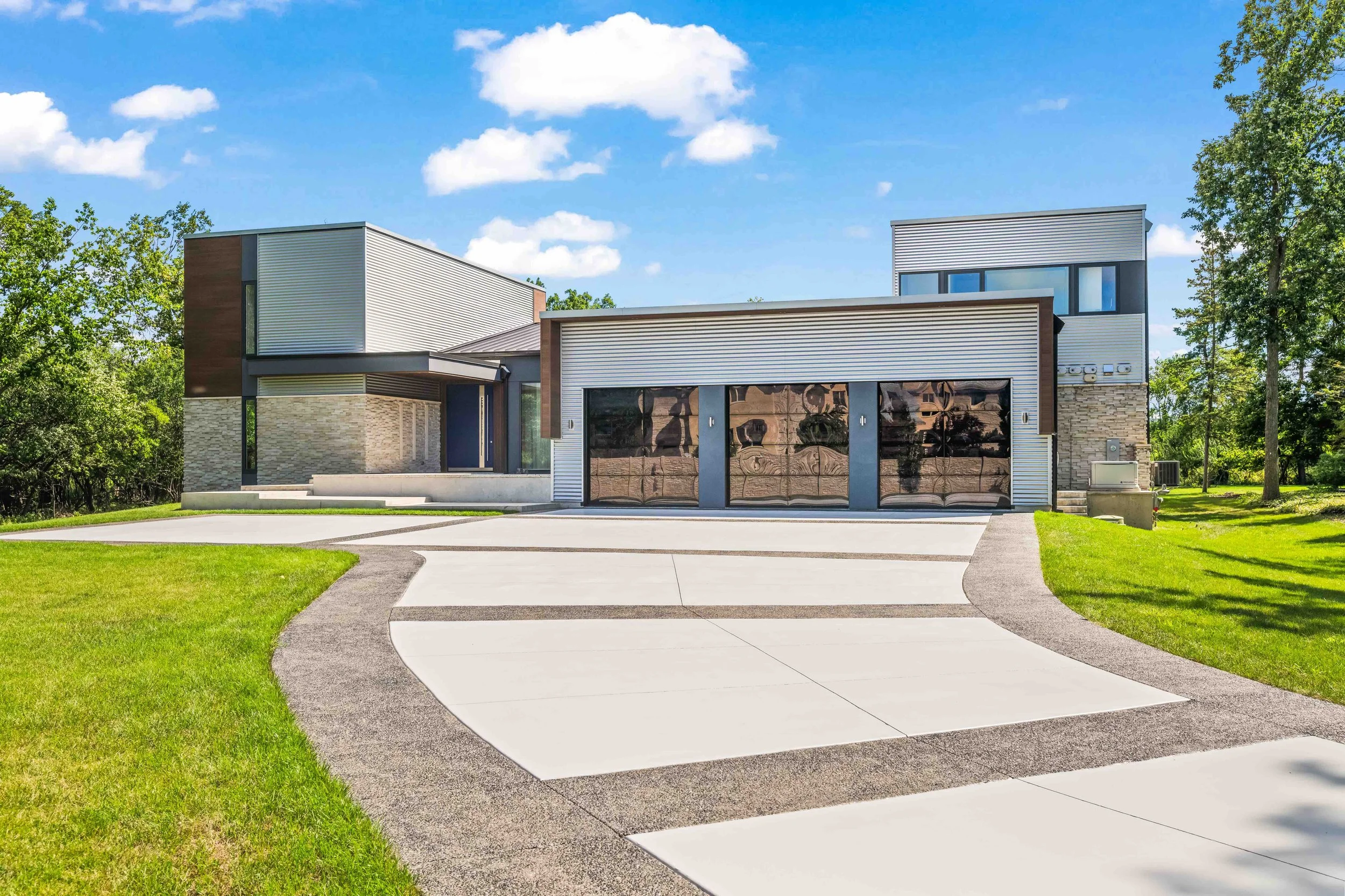 Modern house with flat roof, large driveway with curved edges, green lawn, and blue sky with clouds.