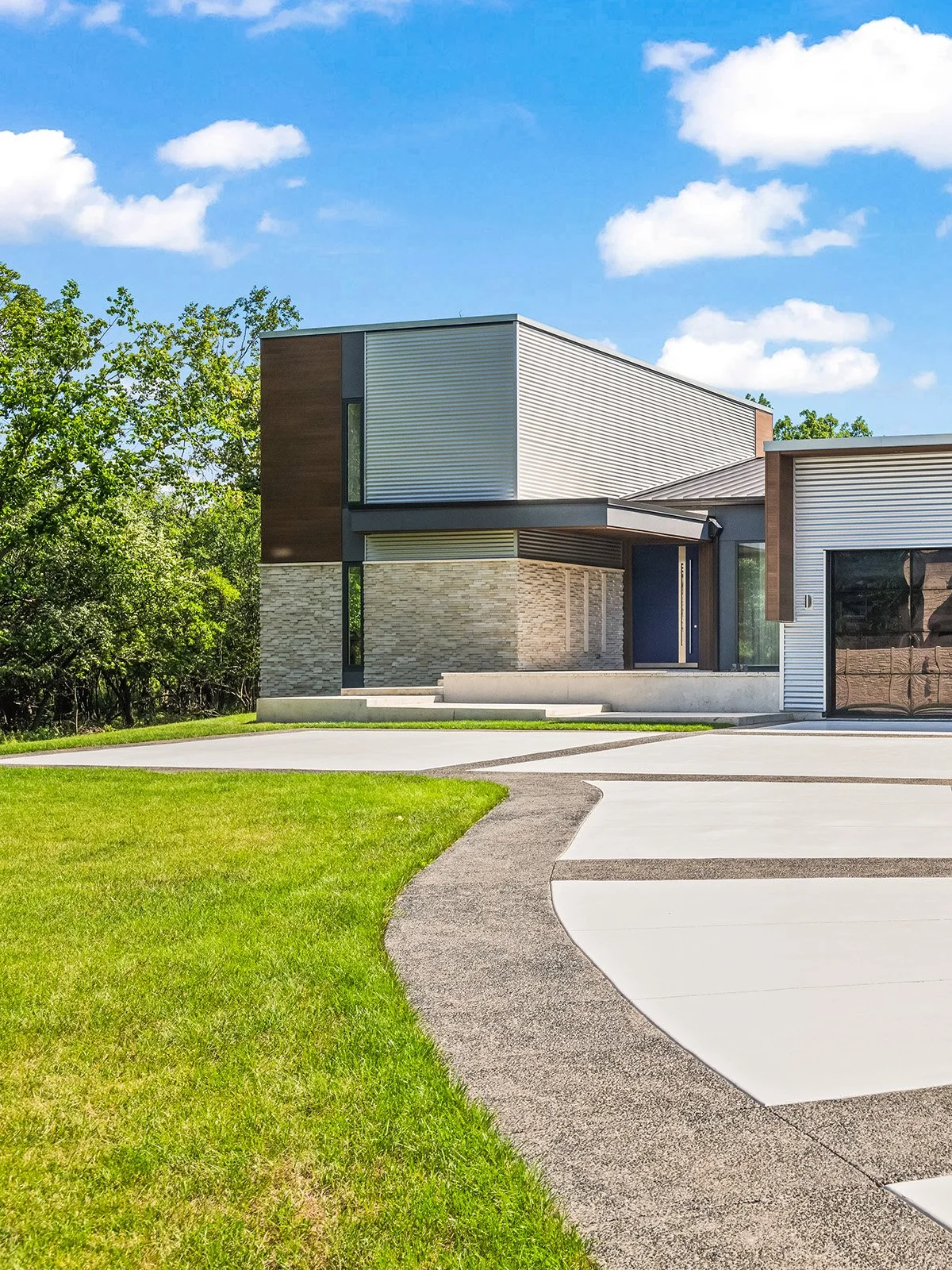 Modern house with a concrete driveway, lawn, and trees in the background under a blue sky with clouds.