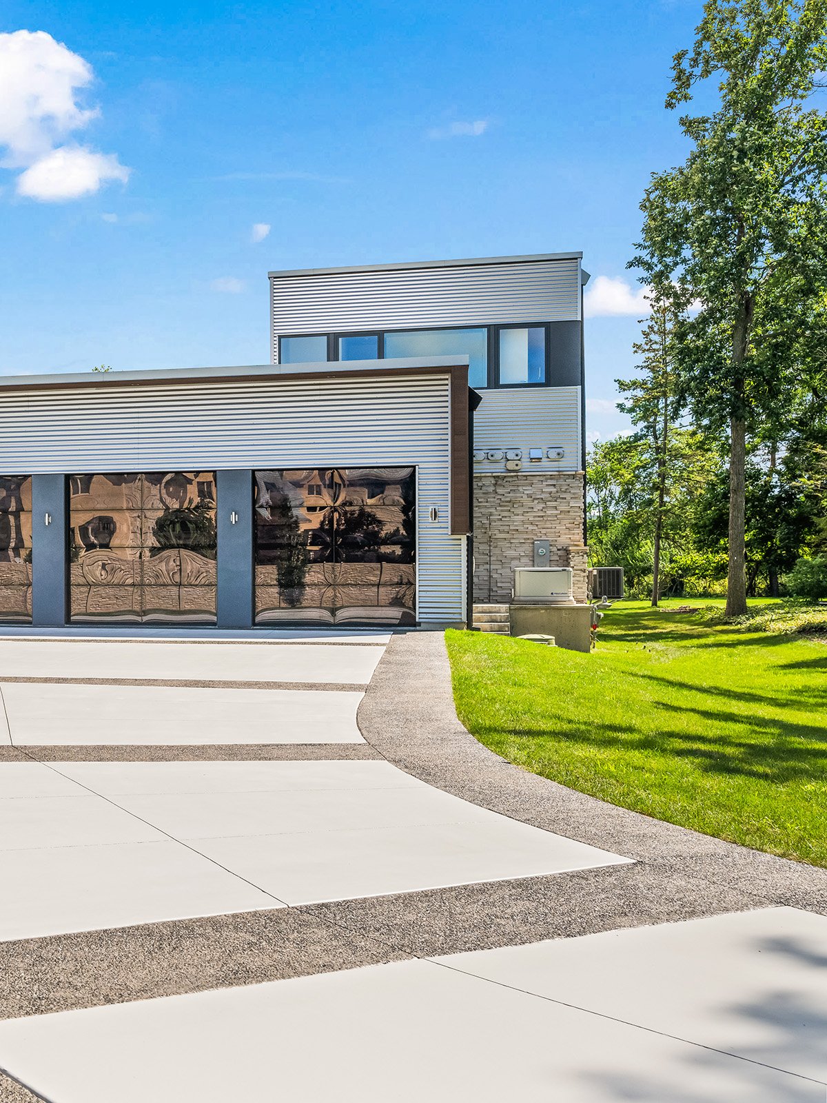 Modern house with a driveway, metal siding, large garage doors with reflective windows, brick accents, surrounded by green grass and trees, under a blue sky with some clouds.