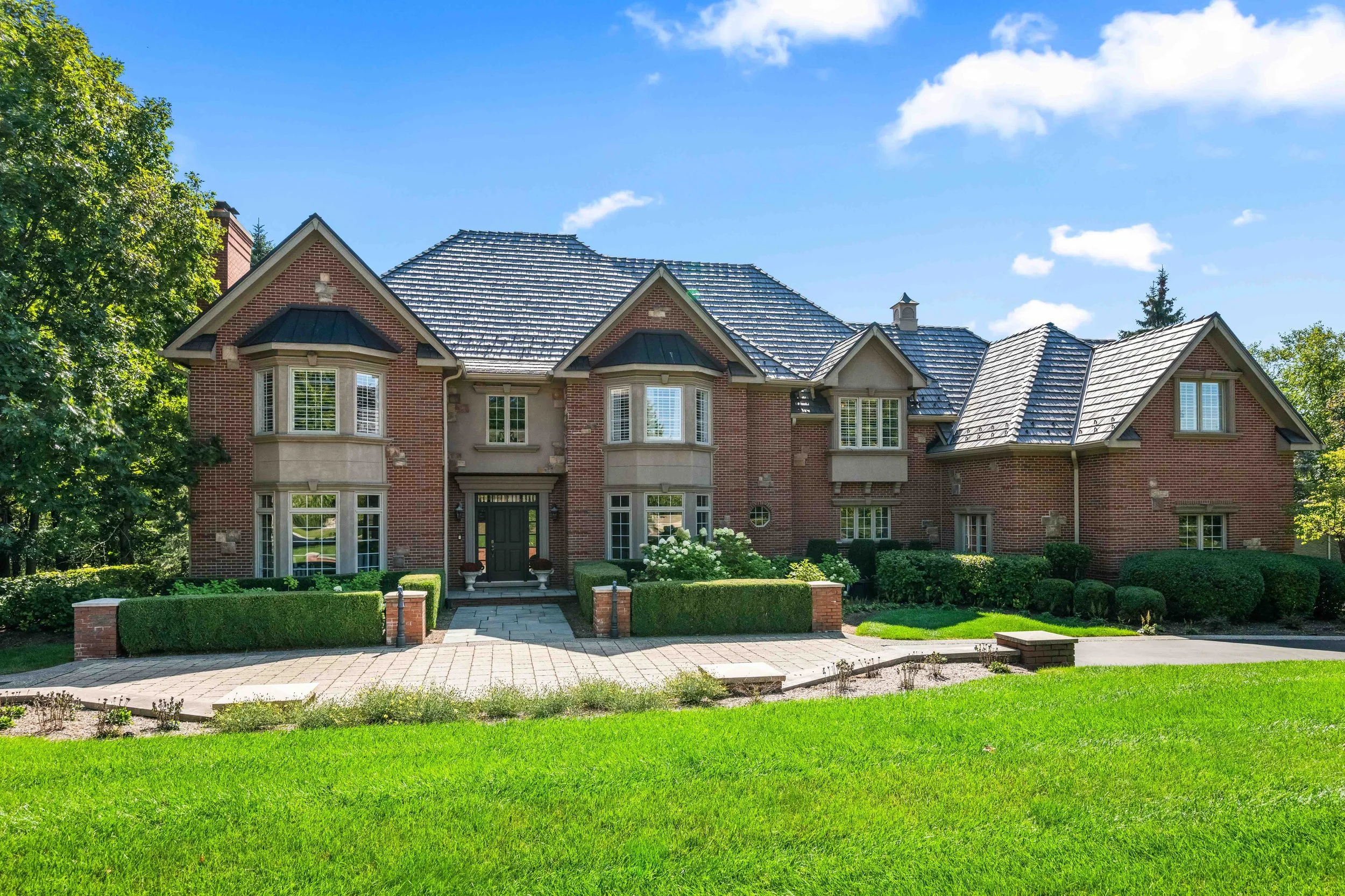 Large brick house with a slate roof, surrounded by a well-maintained lawn and garden, under a partly cloudy blue sky.