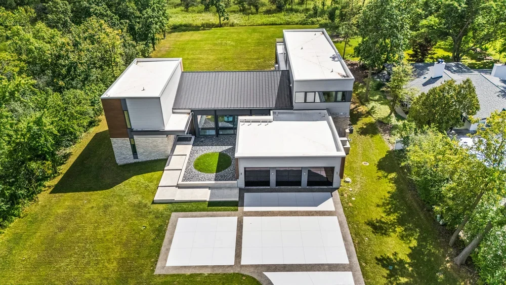 Aerial view of a modern house with flat roofs, large windows, and a well-maintained lawn surrounded by trees.