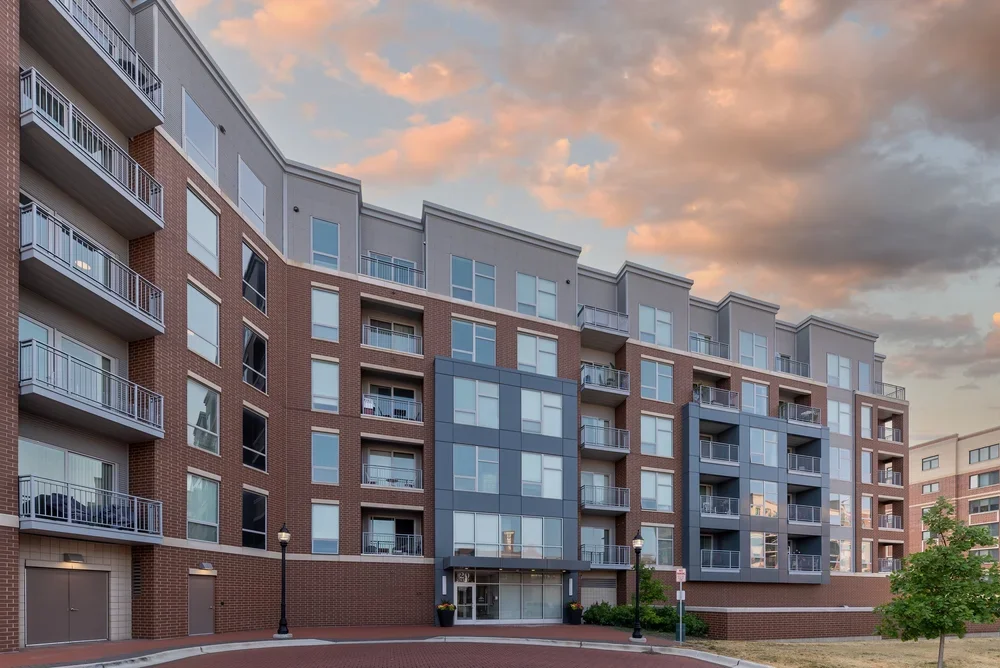 Exterior view of a modern multi-story apartment building with brick and glass facade, several balconies, and a brick-paved driveway at sunset.