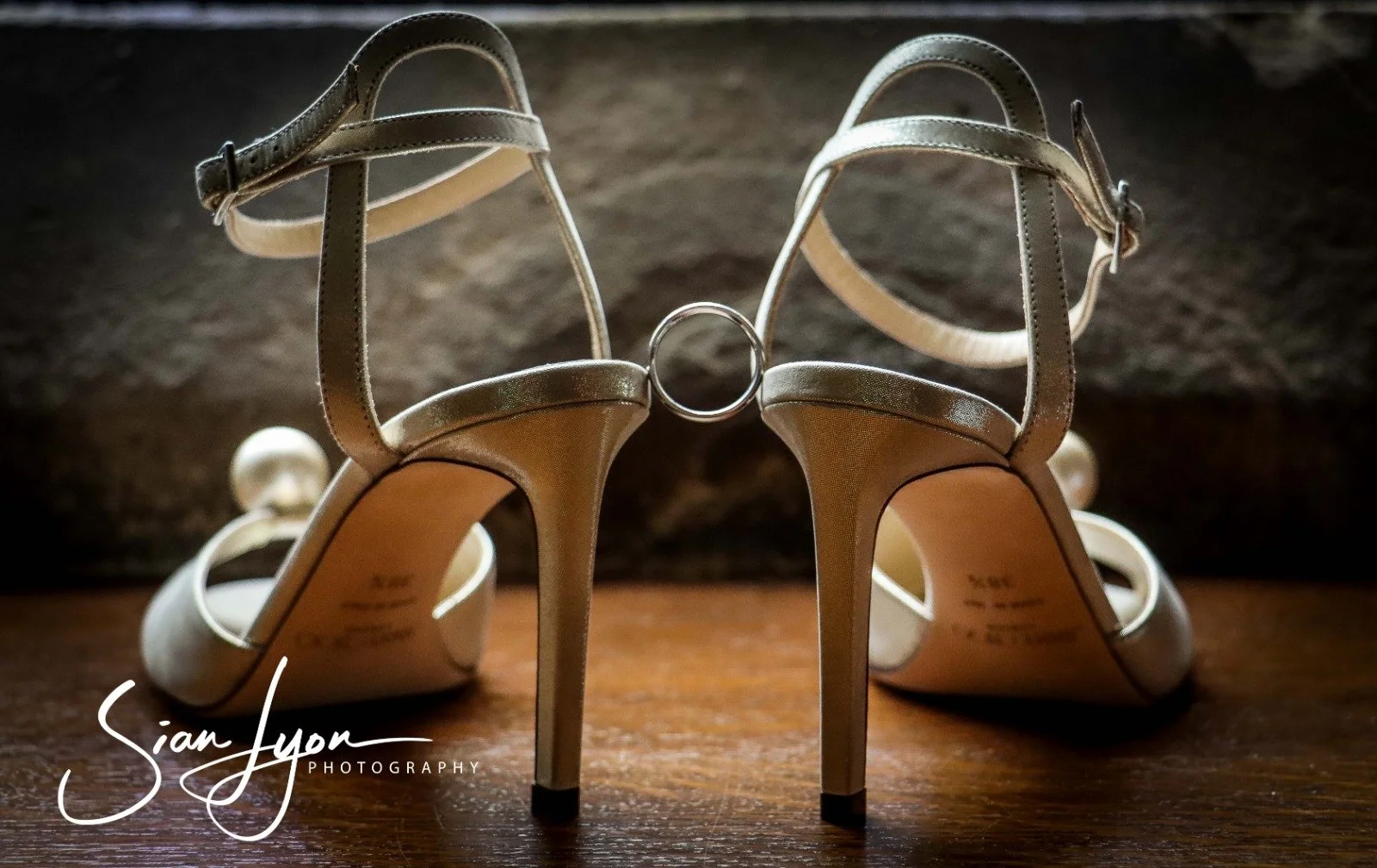 Silver high-heeled sandals with ankle straps and a ring detail between the heels, placed on a wooden surface with a dark background.