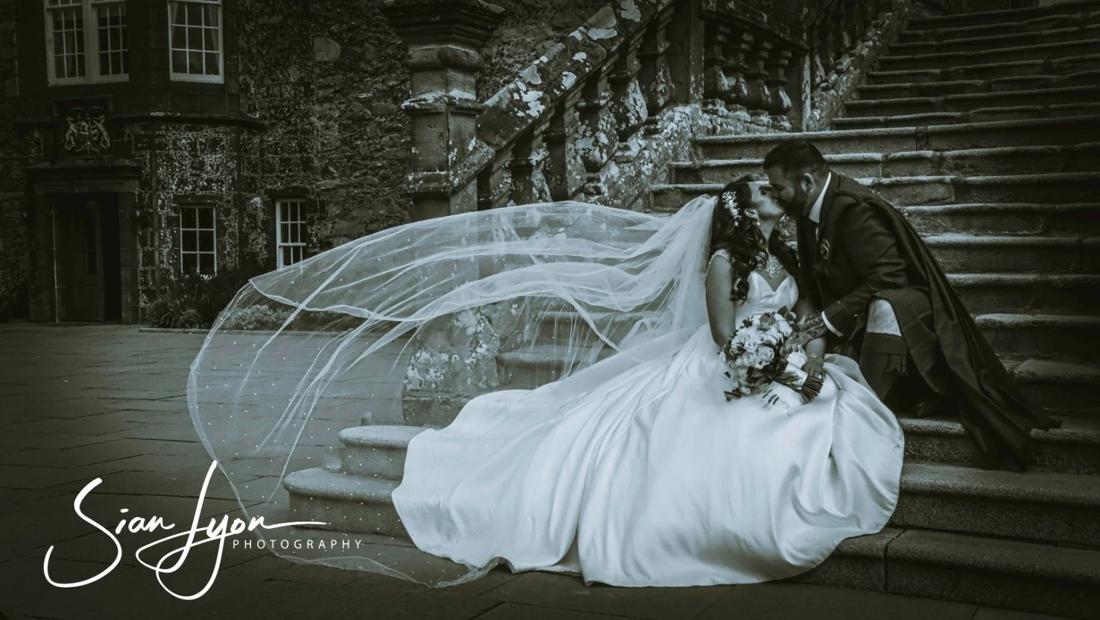 A bride and groom sitting on stone steps outside a historic building, sharing a kiss. The bride is wearing a wedding gown with a long veil and holding a bouquet, while the groom is in traditional Scottish attire including a kilt.