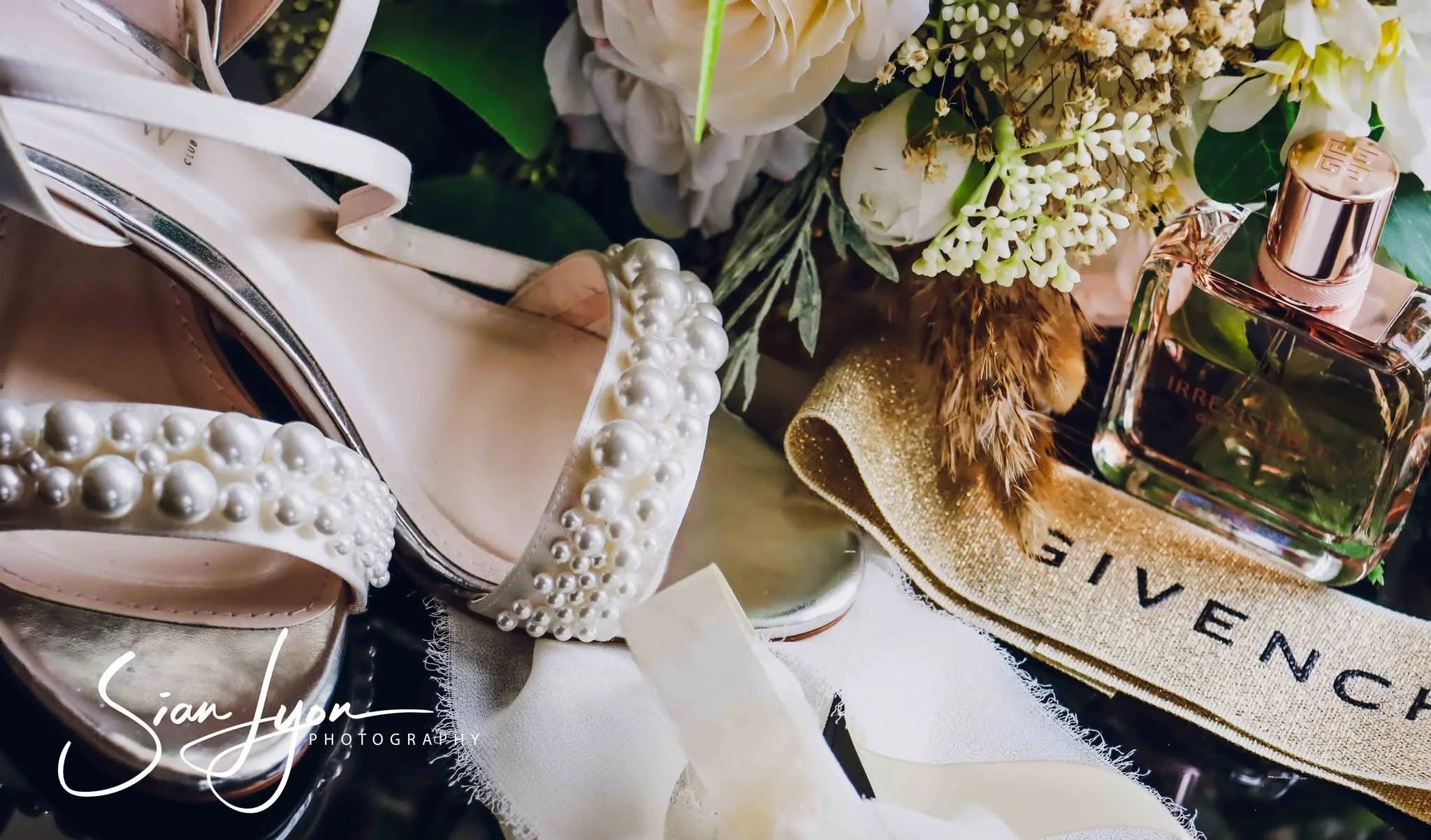 A pair of elegant ivory shoes with pearl embellishments, a bouquet of white and cream flowers, a bottle of Givenchy perfume, and a beige cloth with 'Givenchy' written on it.