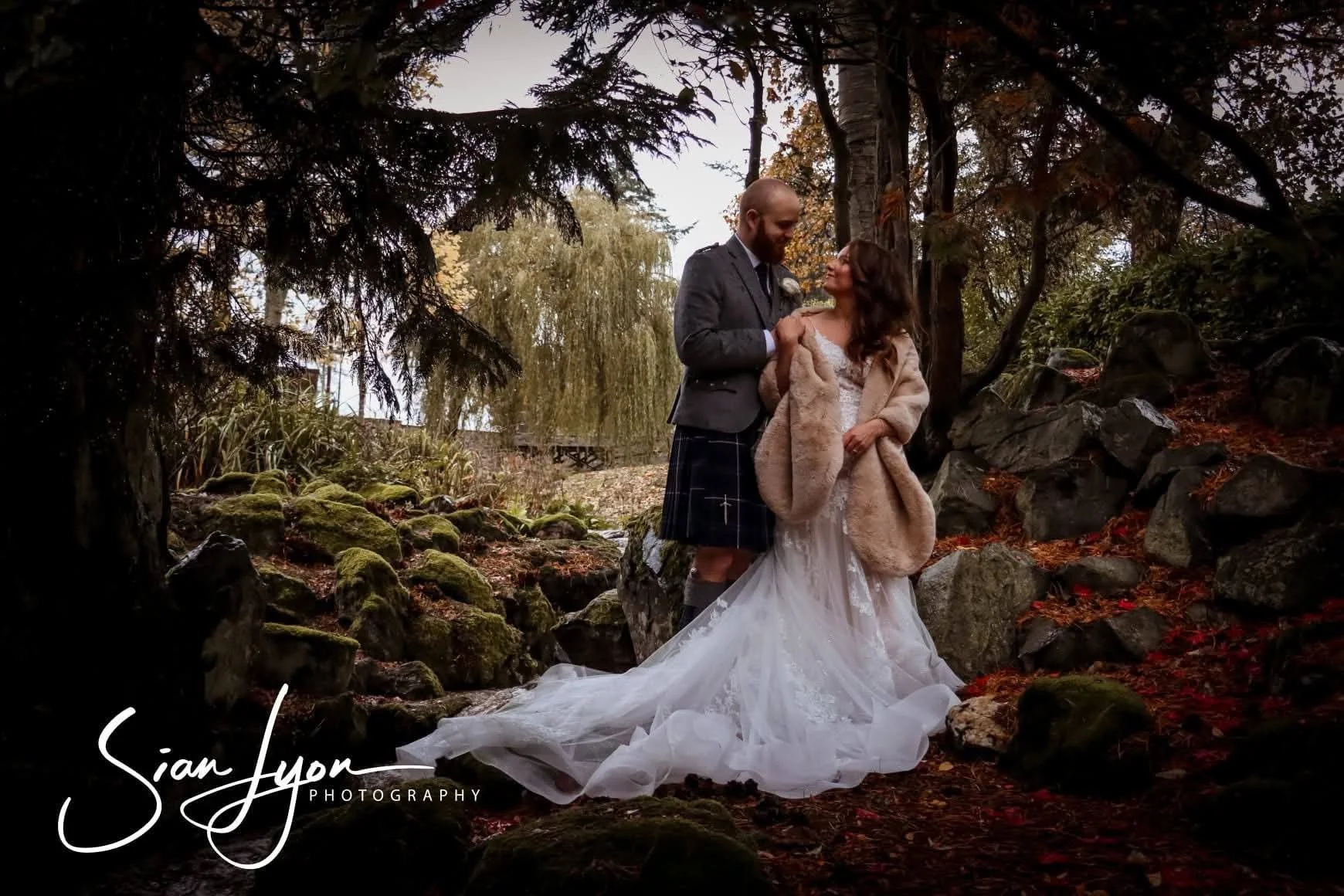 A bride and groom standing together outdoors in a forested area during autumn, holding hands and gazing at each other, with trees, rocks, and fallen leaves surrounding them, and the bride wearing a white wedding dress with a long train and a beige coat. The groom is dressed in a kilt and jacket.
