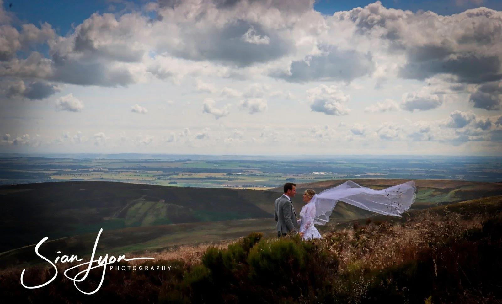 A bride and groom standing on a grassy hillside, holding hands, with a vast landscape and cloudy sky in the background.