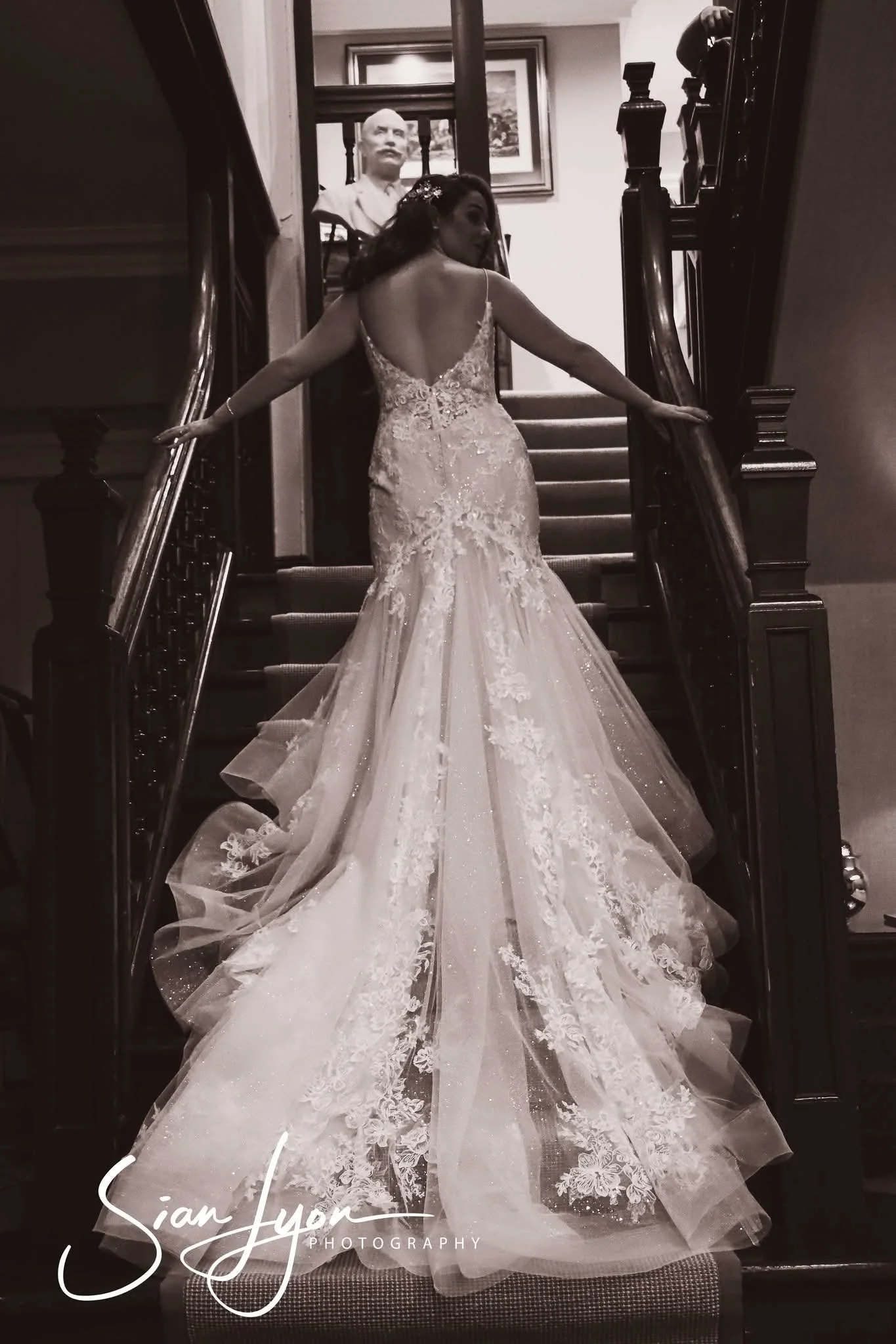 Bride in a wedding gown standing on a staircase with her arms outstretched, facing away from the camera, with a bust sculpture and framed artwork in the background.