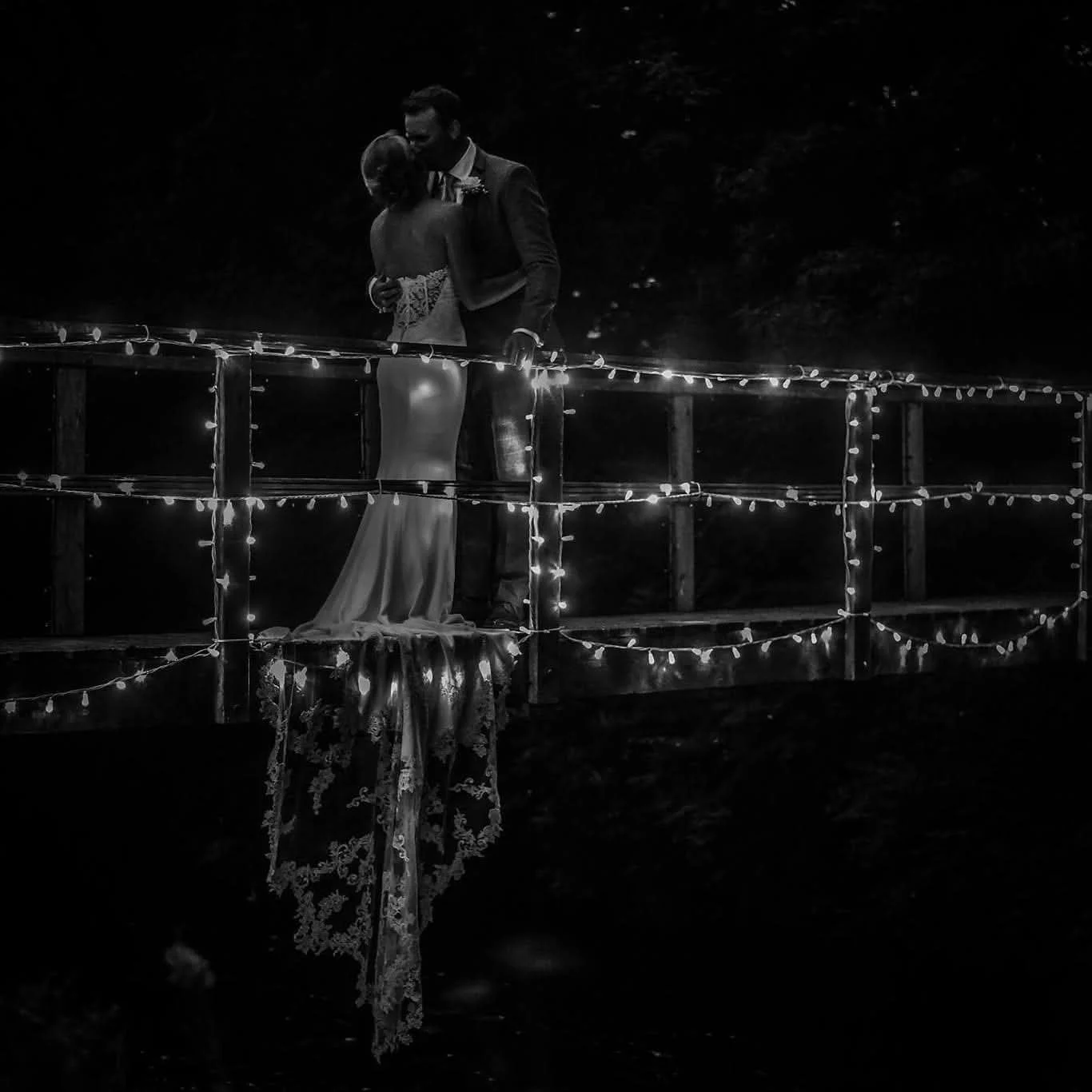 A black and white photo of a couple dancing on a lit-up outdoor bridge at night, with the reflection of a lace wedding dress hanging from the bridge.