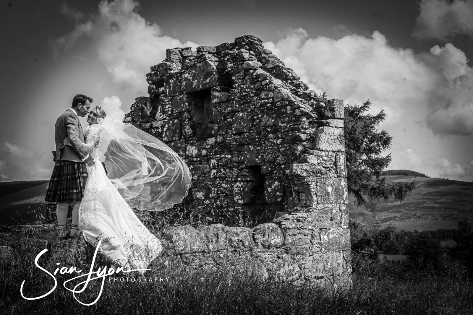 A black and white photograph of a bride and groom standing on grass near a stone ruin. The groom is dressed in a kilt and jacket, and the bride is in a wedding gown with a long veil. They are close together, with the groom holding the bride's waist a