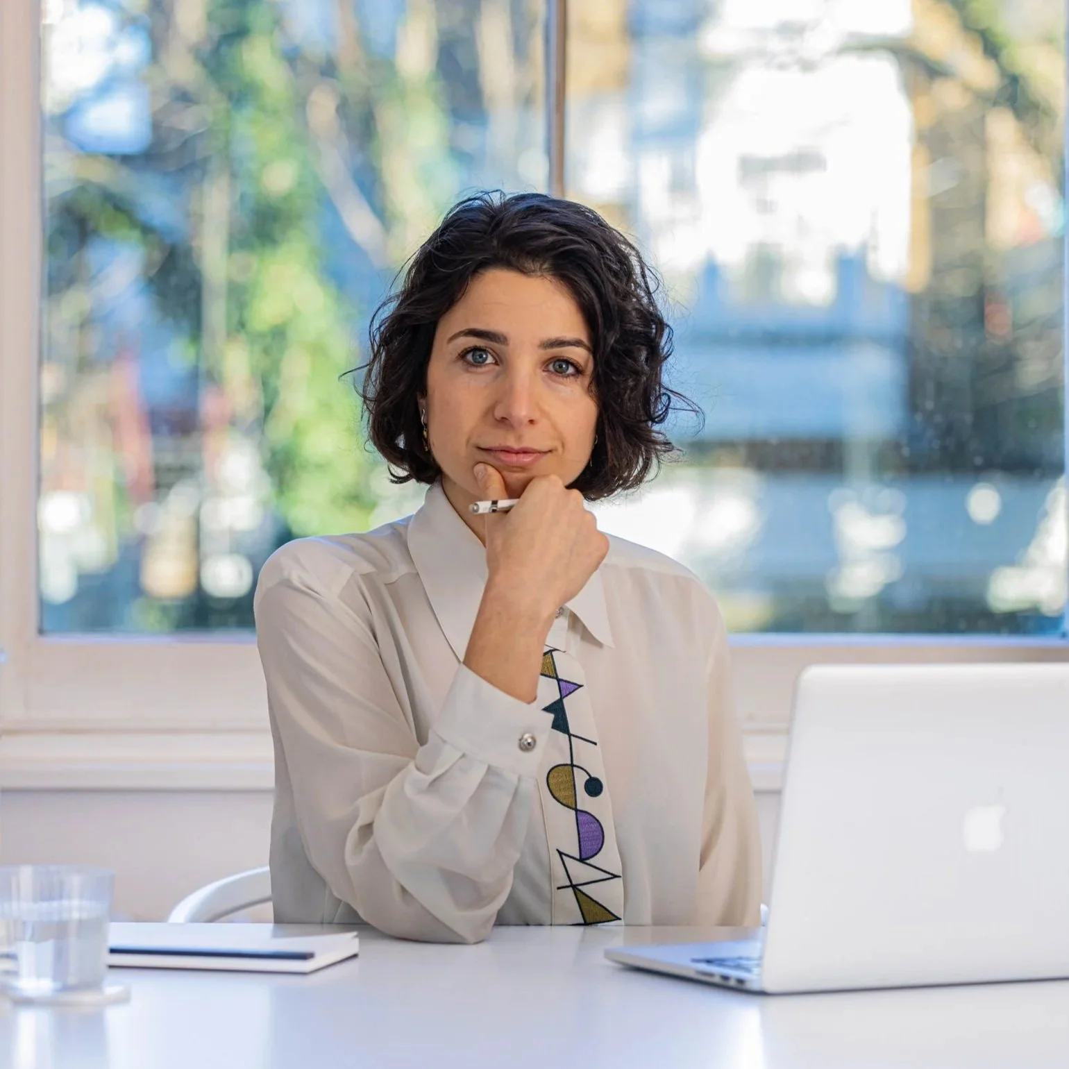 A woman with short, dark, curly hair, wearing a white blouse and a colorful geometric-patterned tie, sitting at a white table with a closed notebook, a glass of water, and a laptop, in front of a window with a blurred outdoor scene.