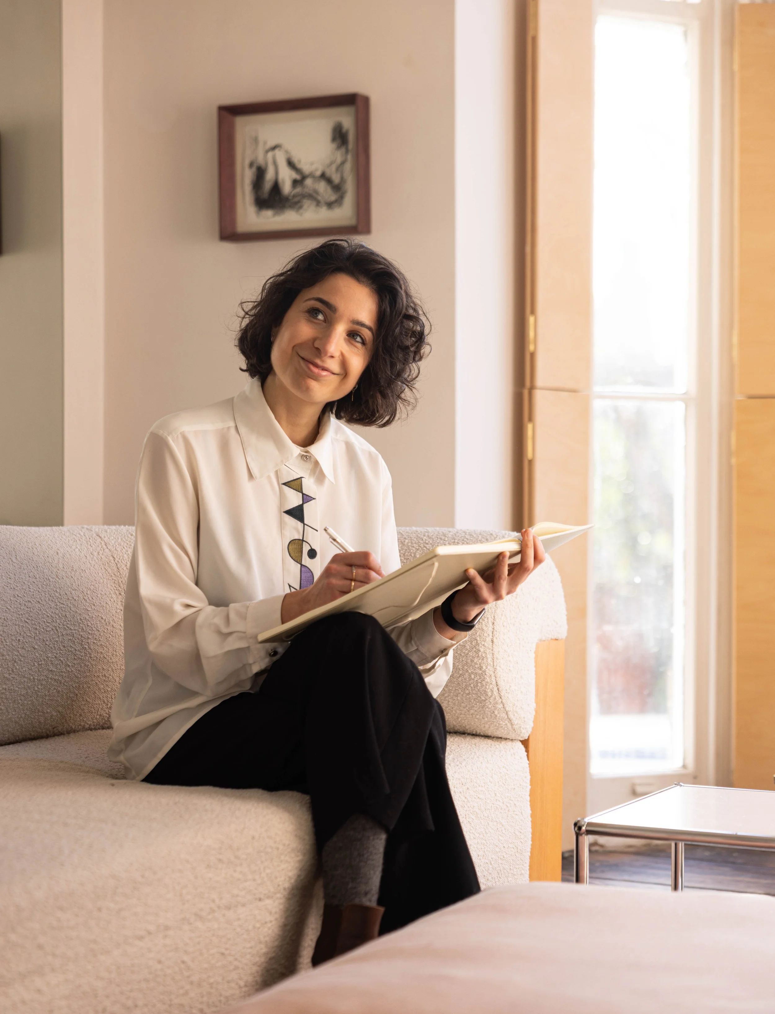 A woman with dark curly hair sitting on a cream-colored couch, smiling, holding a notebook and pen indoors near a window in a well-lit room.
