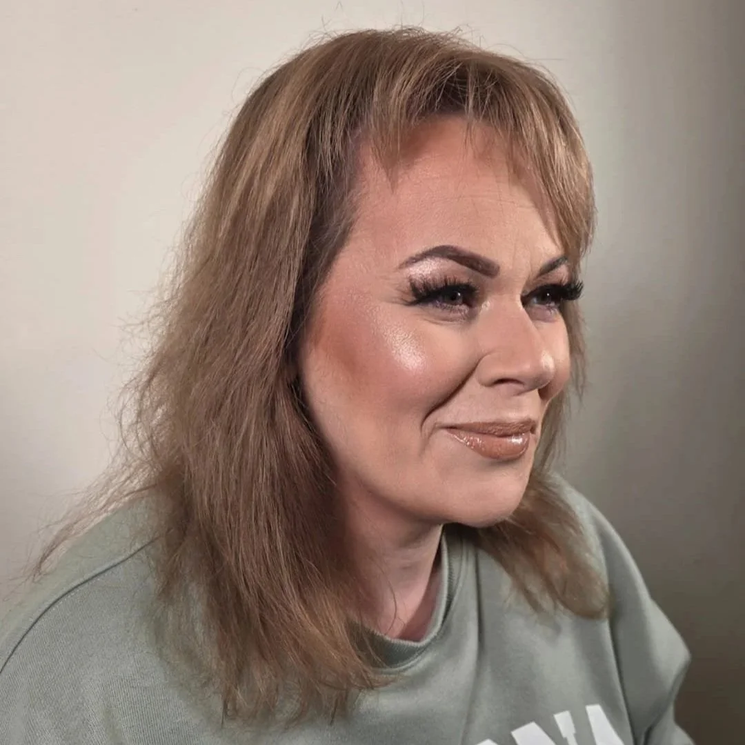 A woman with light brown, shoulder-length hair and makeup is smiling slightly against a plain background.