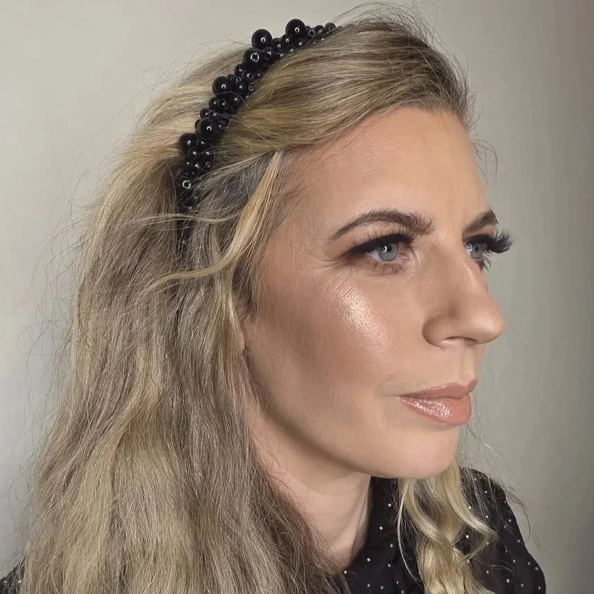 A woman with blonde hair wearing a black beaded headband, makeup, and a black polka dot top, looking to her right.