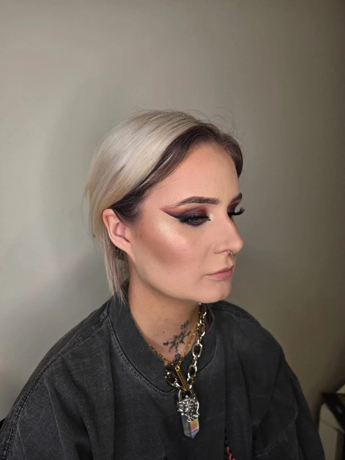 A woman with platinum blonde and dark brown hair, wearing dramatic eyeliner and makeup, a black jacket, and layered necklaces, looking to the side against a plain background.