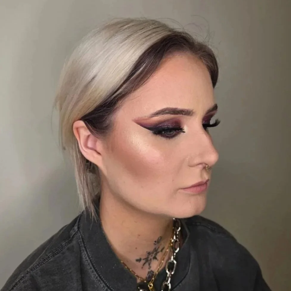 A woman with short, platinum blonde hair, wearing dark makeup with bold eyeliner and eyeshadow, multiple necklaces, and a dark shirt.