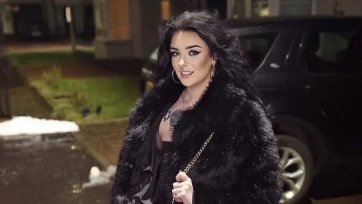 A woman with long black hair, dressed in a black outfit, wearing a black fur coat, holding a small purse with a chain strap, standing outdoors at night in front of a black vehicle.