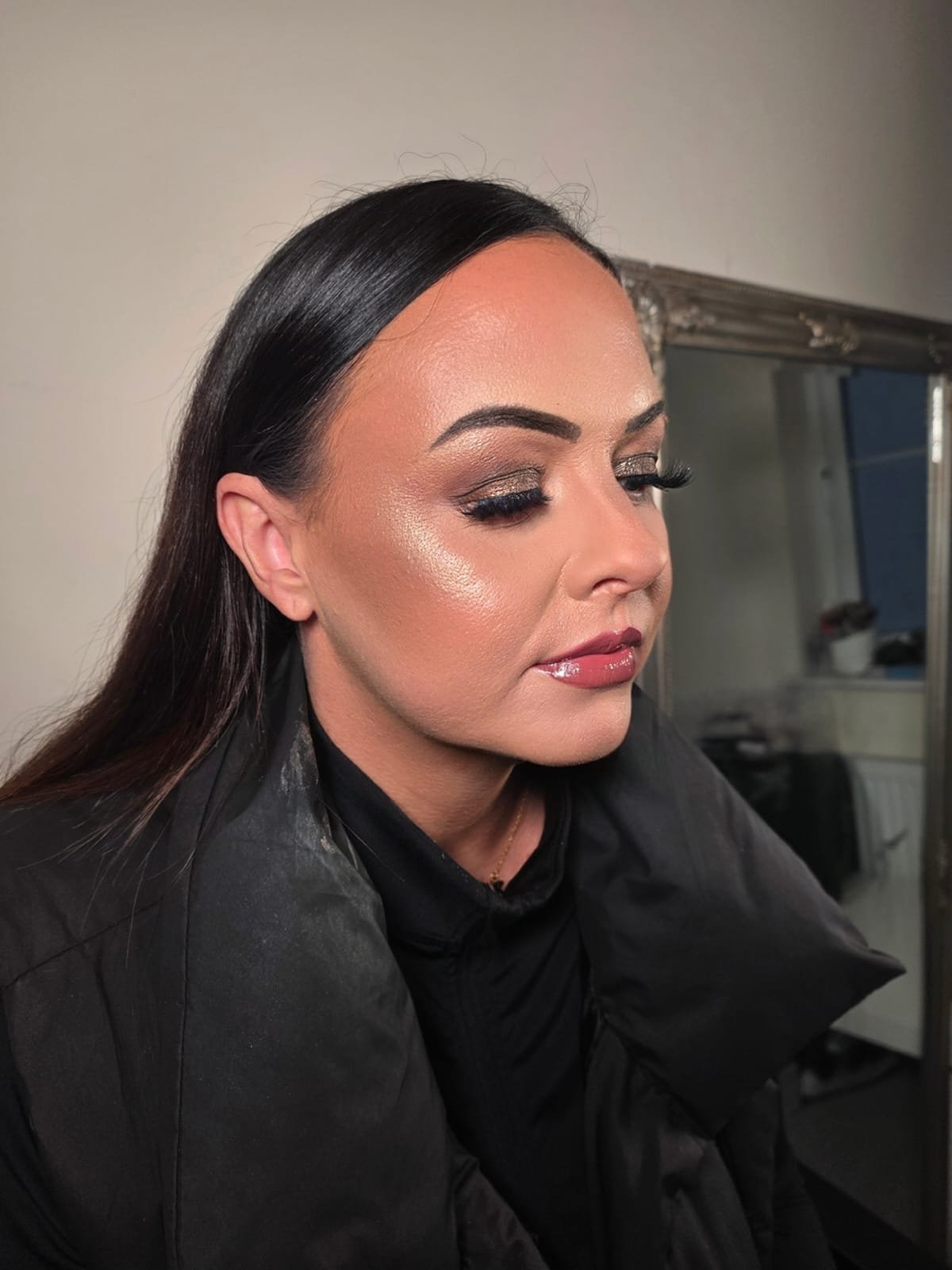 Close-up of a woman with dark hair and makeup, including eyeshadow, mascara, highlighter, and glossy lips, sitting in a makeup chair.