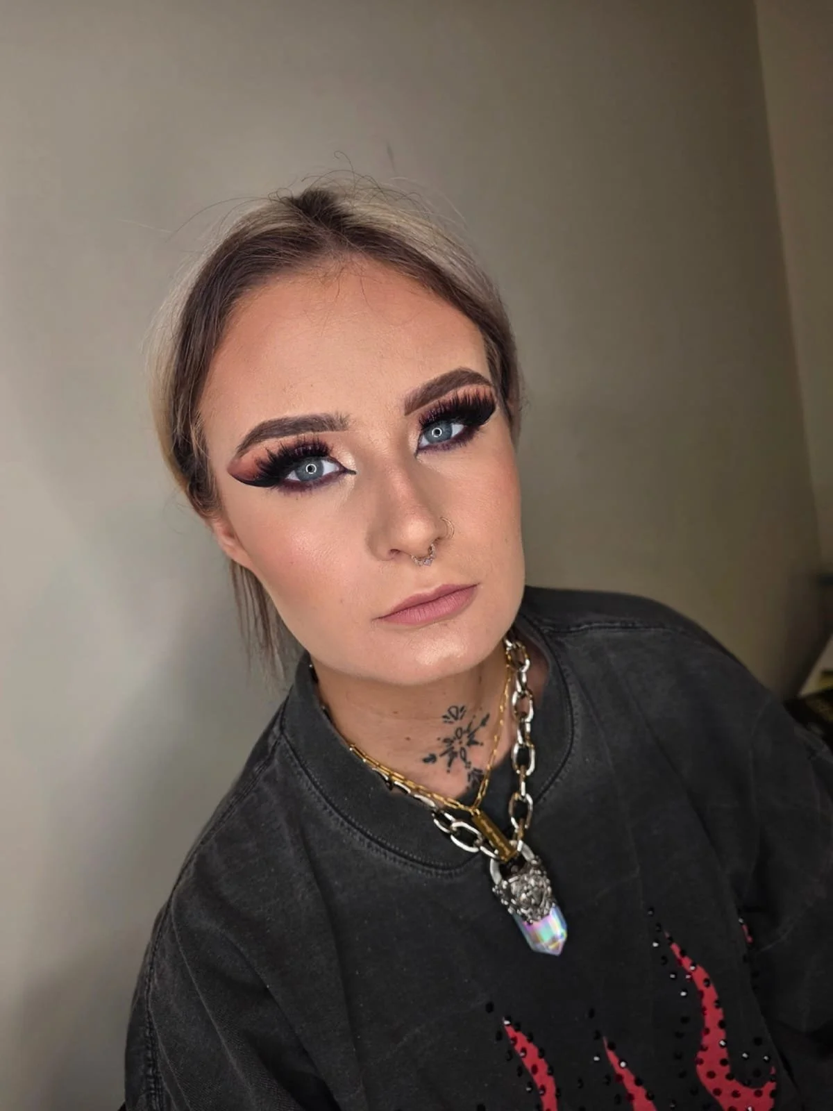 A woman with platinum blonde hair, dramatic makeup, and blue eyes wears a black denim jacket, layered necklaces, and has a septum piercing.