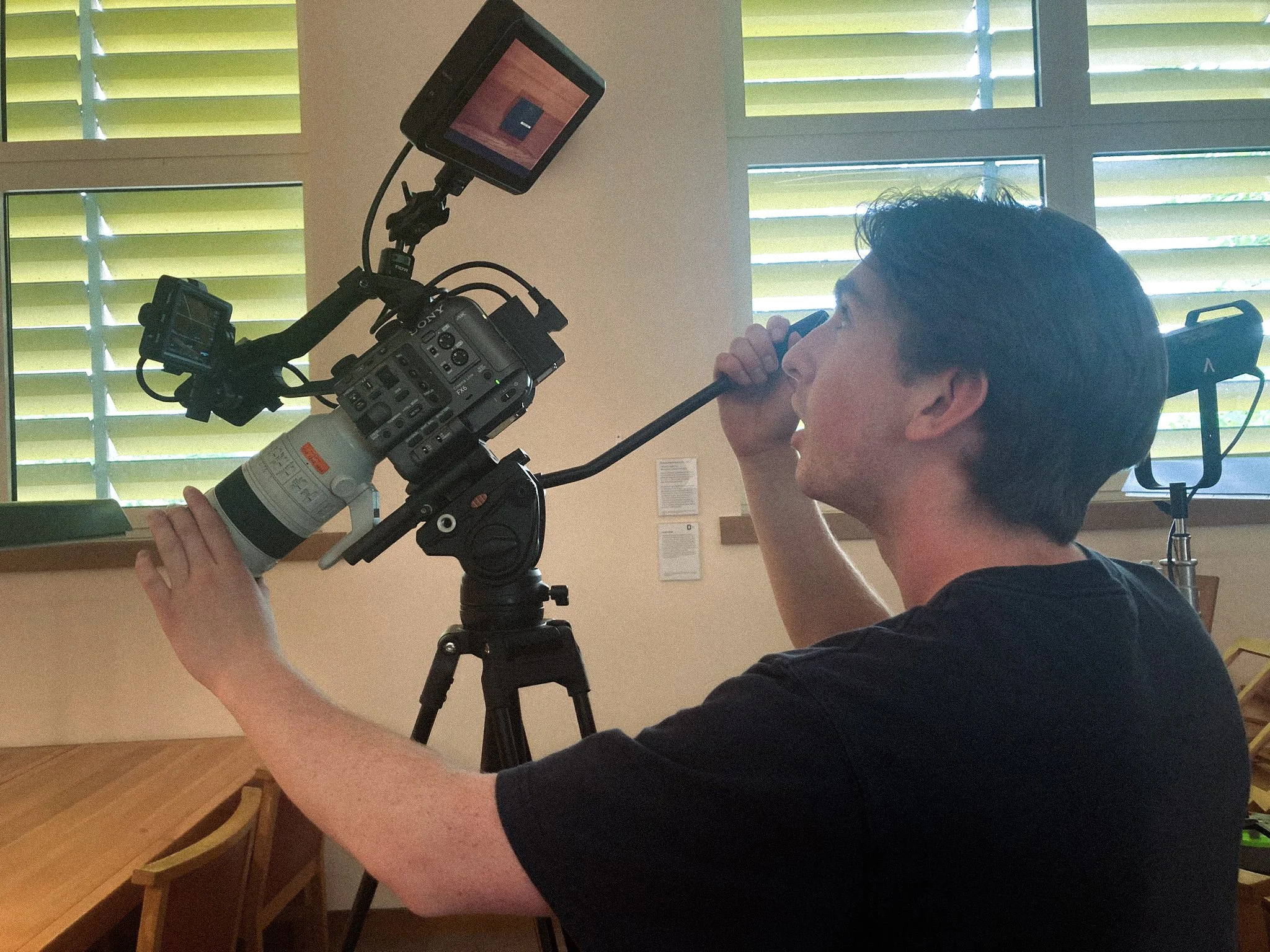 A man operating a video camera on a tripod, drinking a beverage through a straw, with green window blinds in the background.