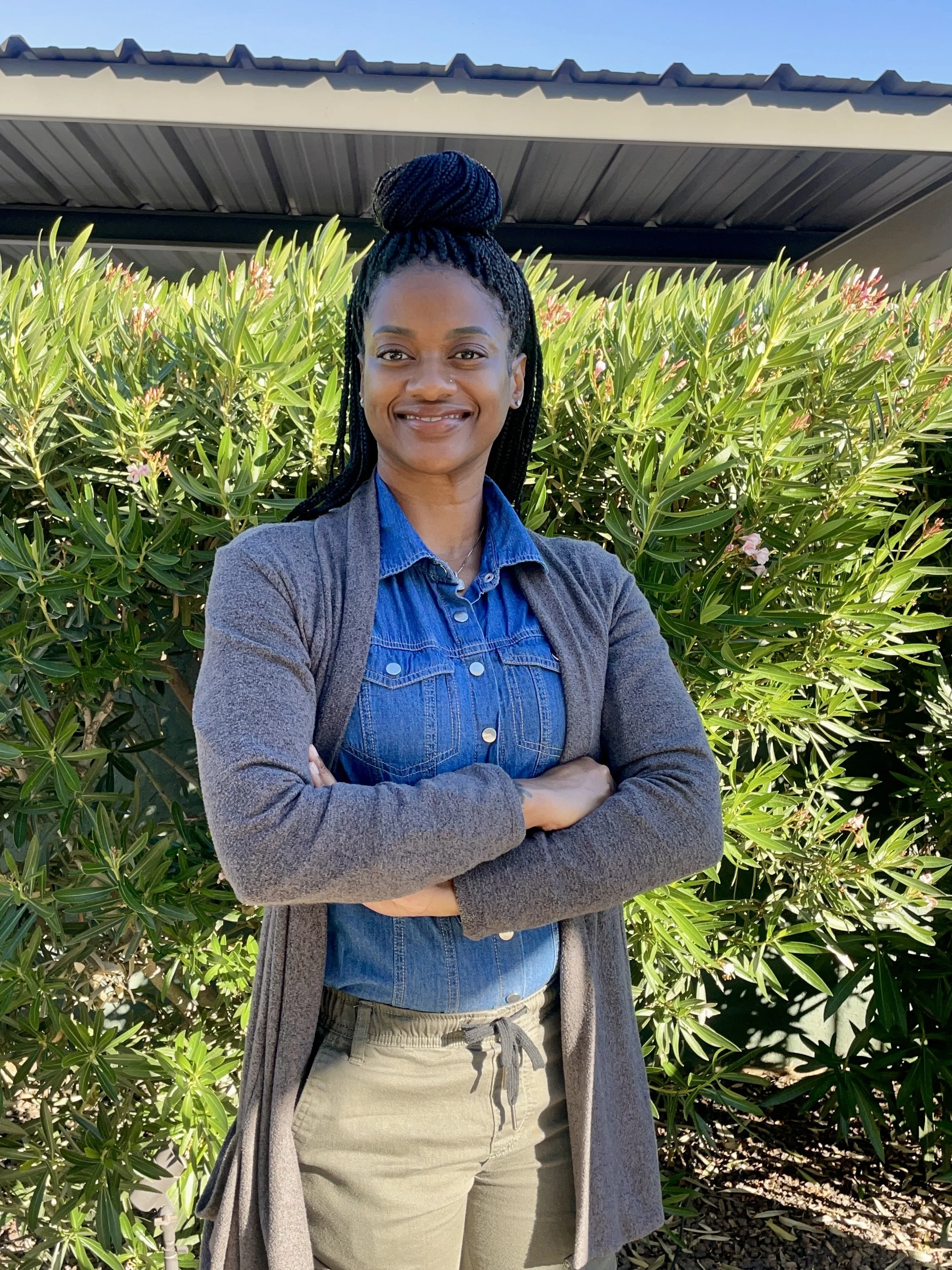 A smiling woman with braided hair styled in a top bun, standing outdoors in front of green shrubbery under a clear sky.