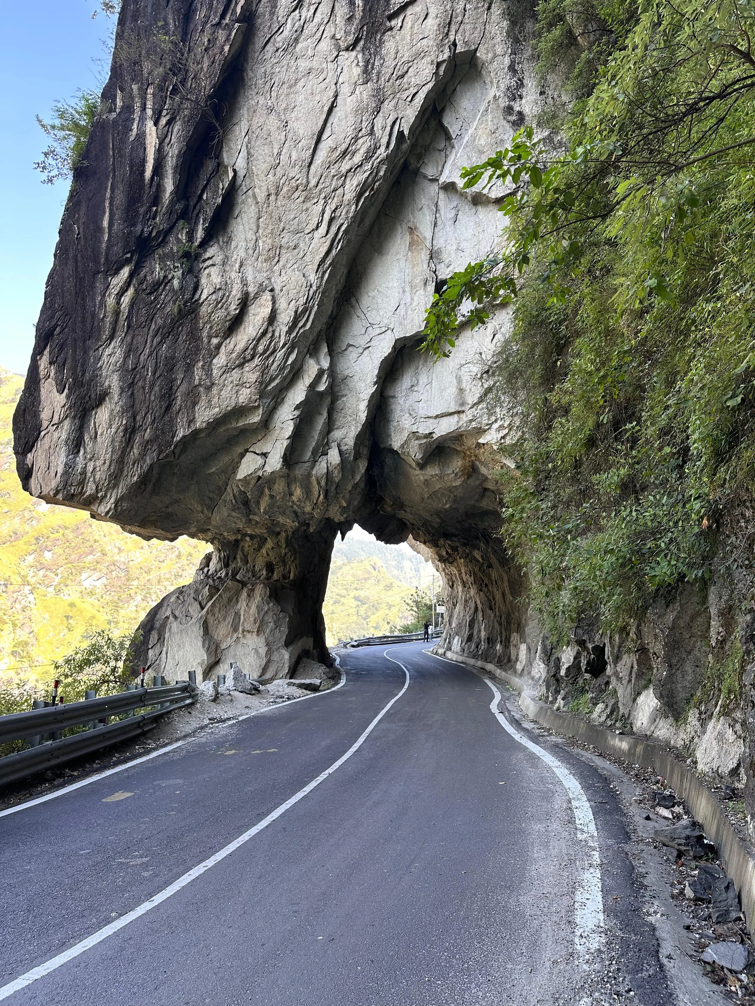 A winding mountain road in Kinnaur passing through a large rock formation that creates a natural tunnel, with lush green trees on the right side.