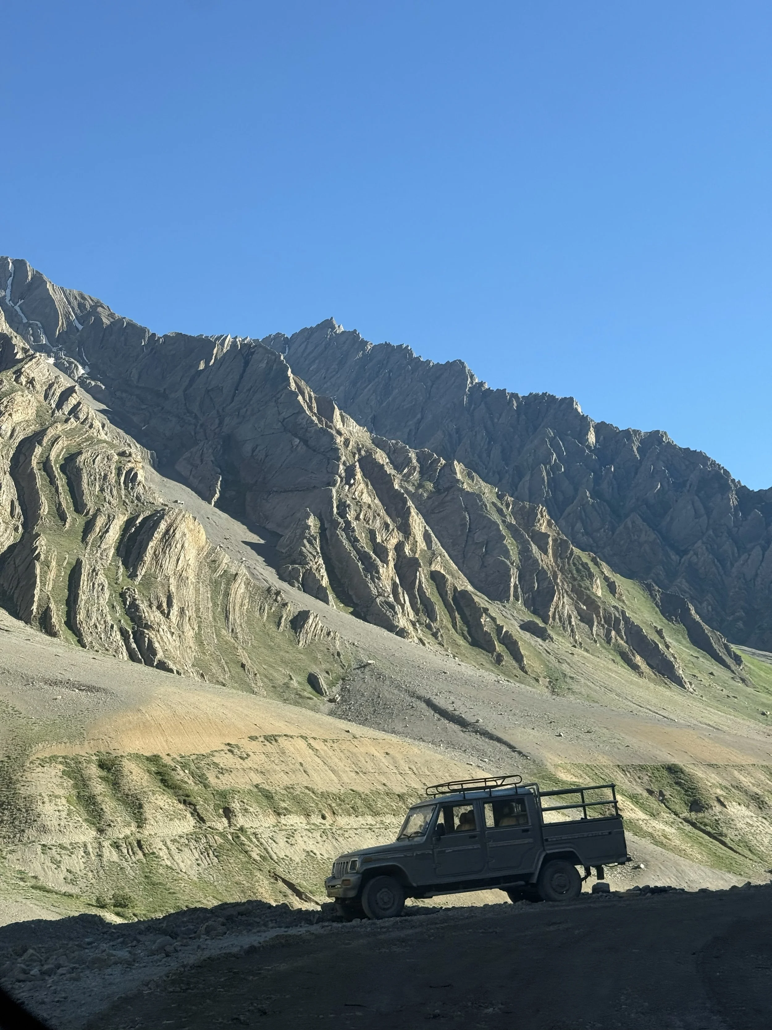 A rugged black pickup truck parked on a dirt road in front of a mountain range with steep, rocky slopes and a clear blue sky.