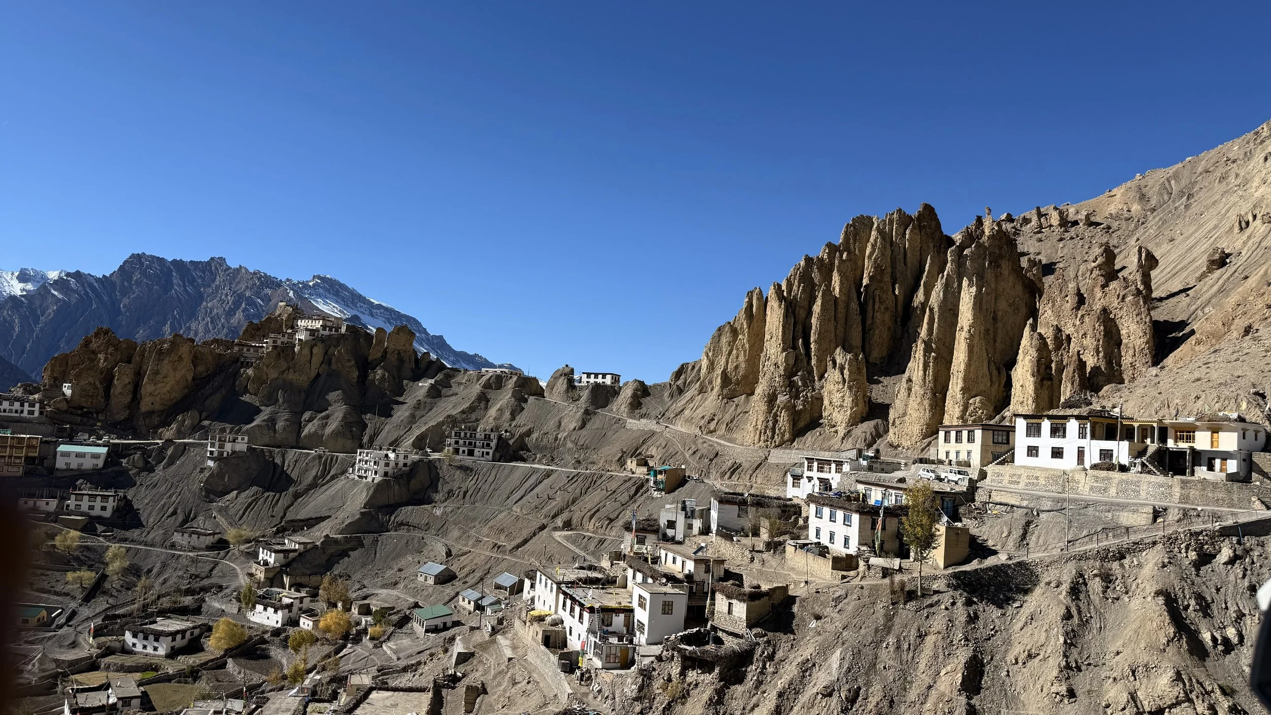 A Spiti village set against steep rocky cliffs, with scattered white homes and distant snow-capped peaks under clear skies
