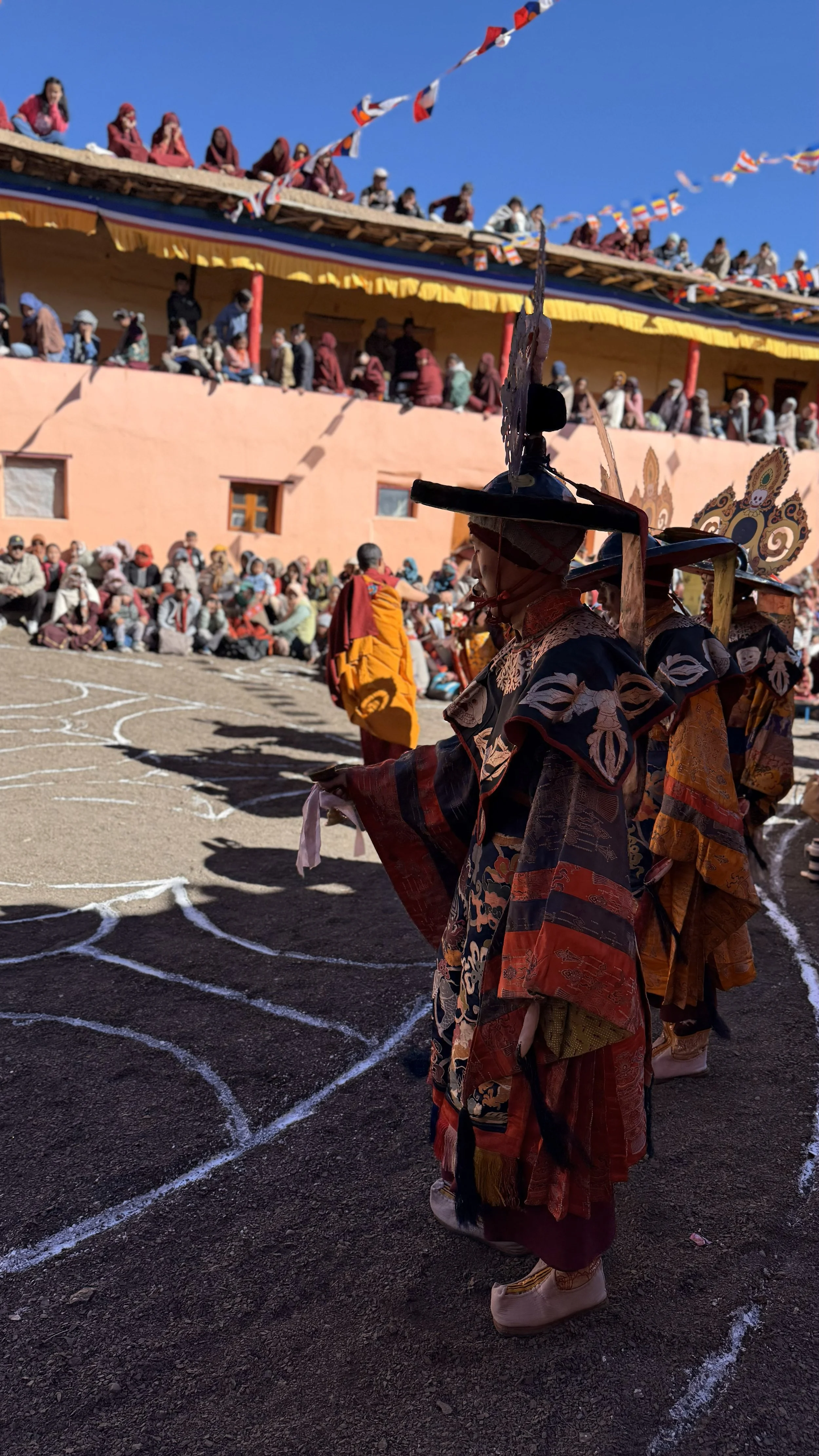 Monks in ornate robes participating in a cultural dance or ceremony at a temple, with spectators watching from the stands and a clear blue sky overhead.