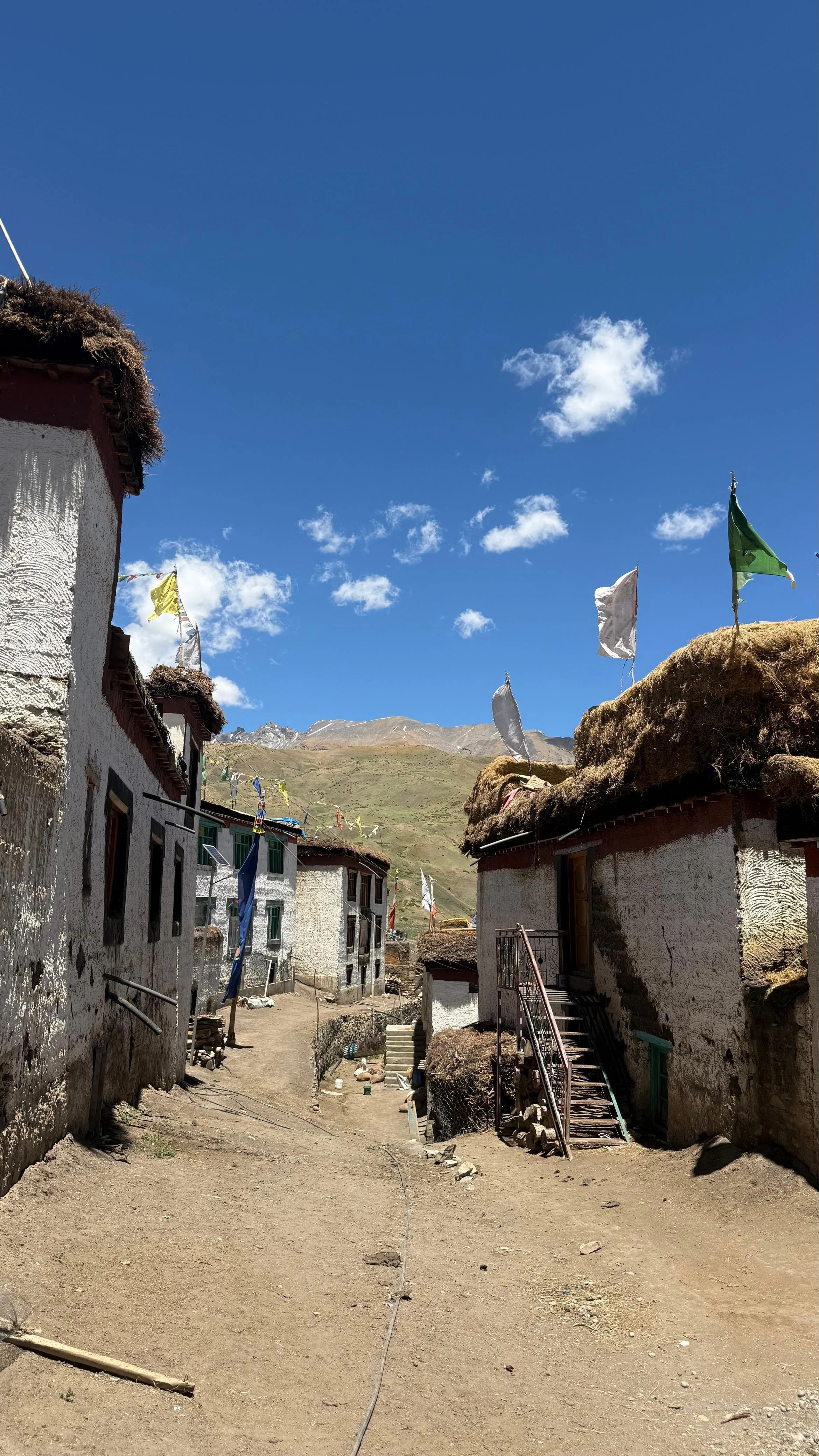 High-altitude Spiti village with traditional mud-brick houses, flat roofs, narrow footpaths, prayer flags, and barren brown mountains under a wide blue sky.
