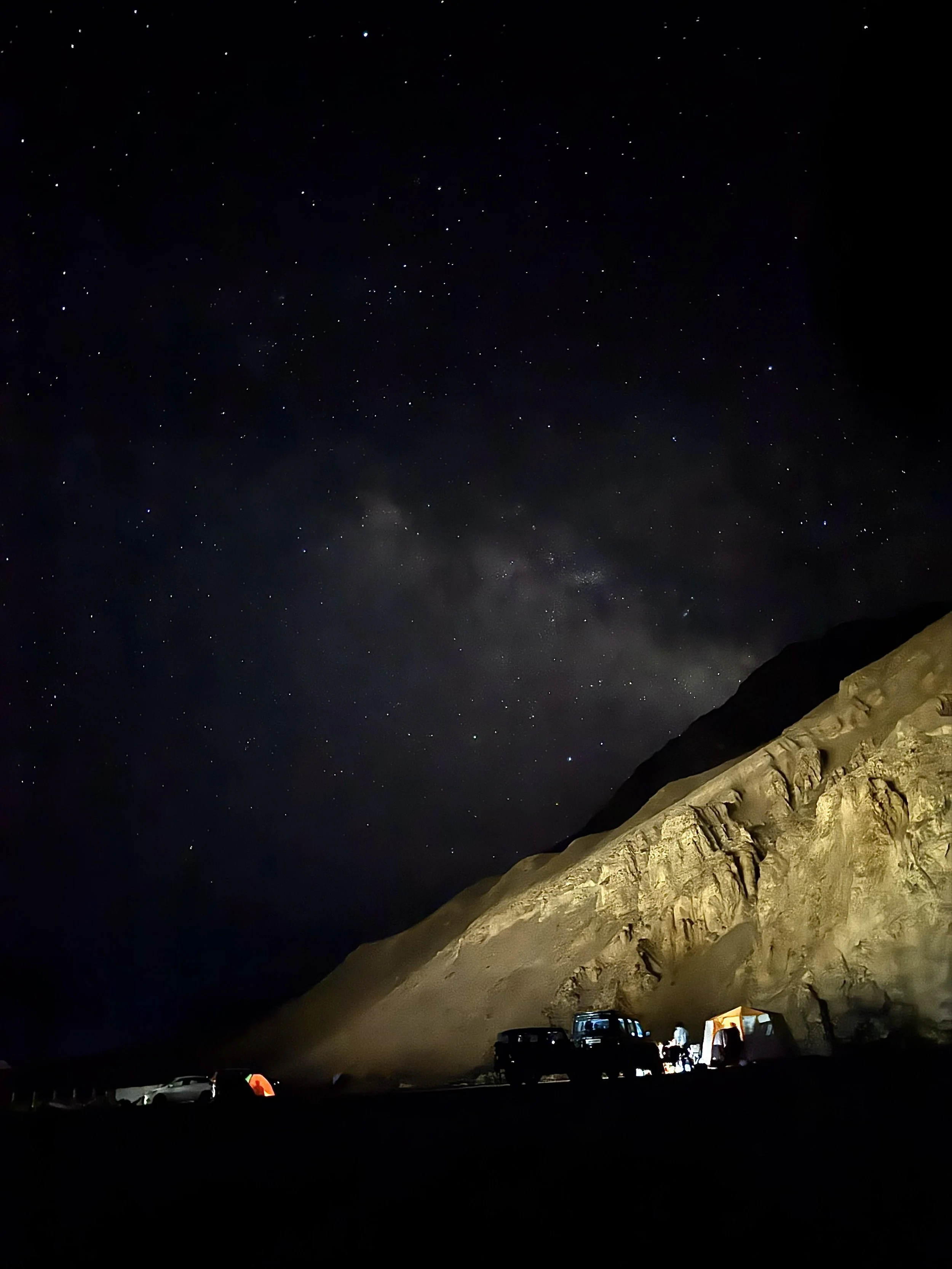 Night sky filled with stars above a rugged hillside with tents and parked cars at a campsite. Beautiful Himachal