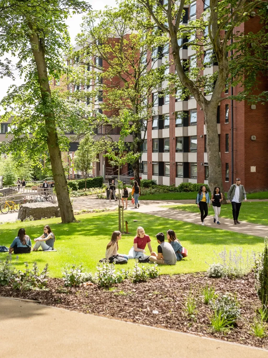 People sitting on the grass and walking in a park area near a red-brick apartment building with green trees and pathways.