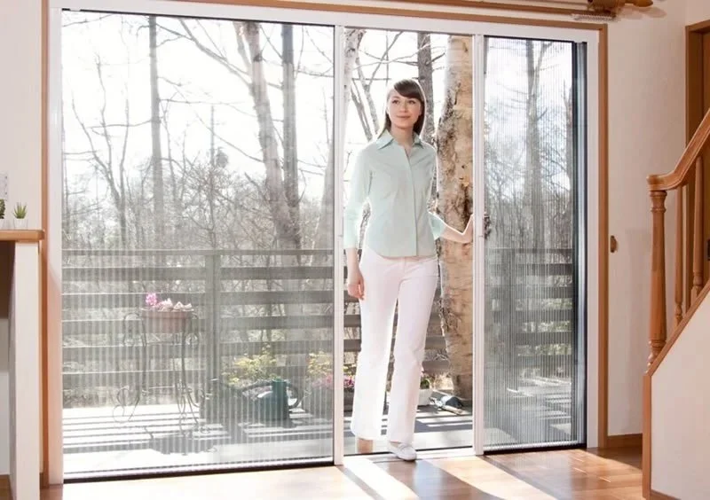 Woman standing at a glass sliding door in a bright room, holding the door open to a backyard with trees and potted plants.