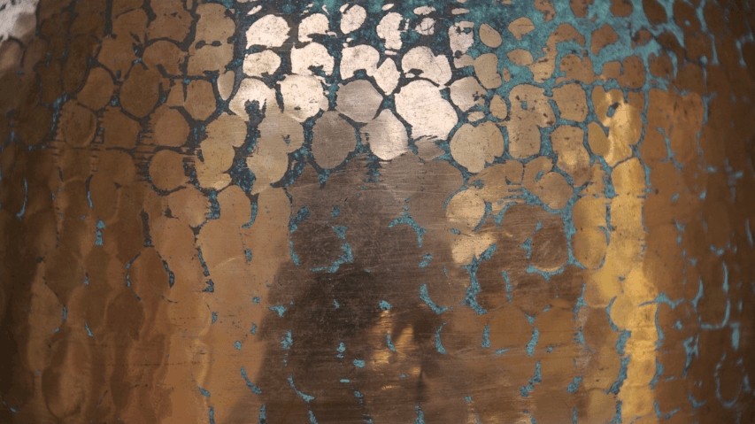 Close-up of a metallic surface with a pattern of hexagonal shapes and a central figure that resembles a person, in shades of copper, with some areas showing signs of oxidation or patina.