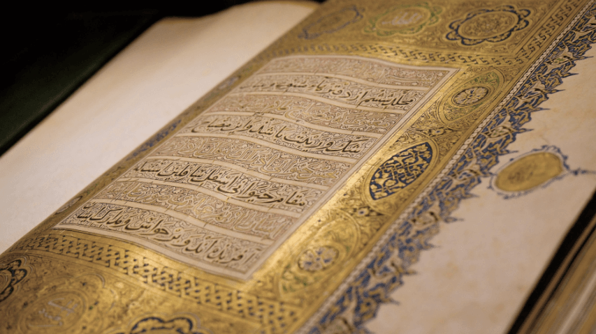An open, ancient manuscript with intricate gold, blue, and red decorative borders and Arabic calligraphy on cream-colored pages.