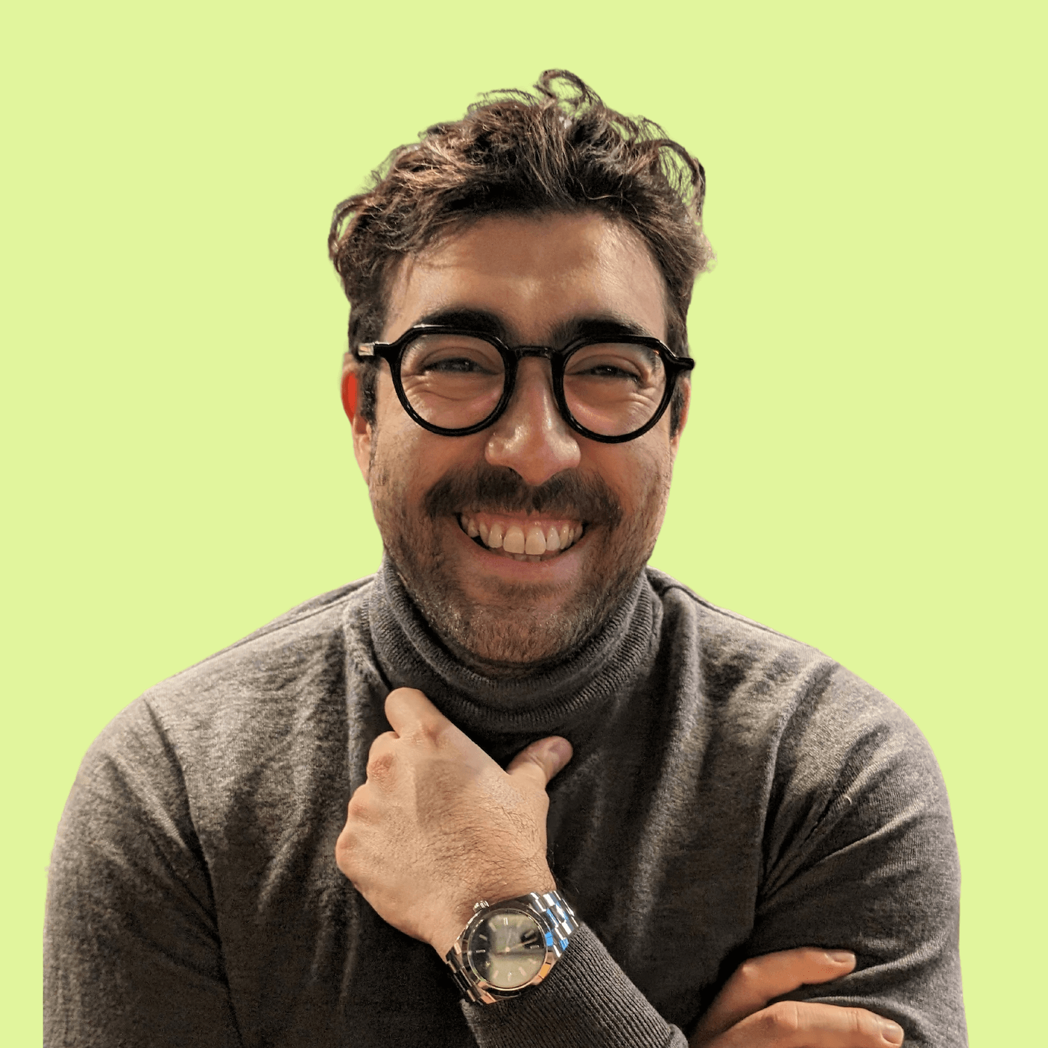 A smiling man with glasses and a beard, wearing a gray turtleneck and a watch, against a light green background.