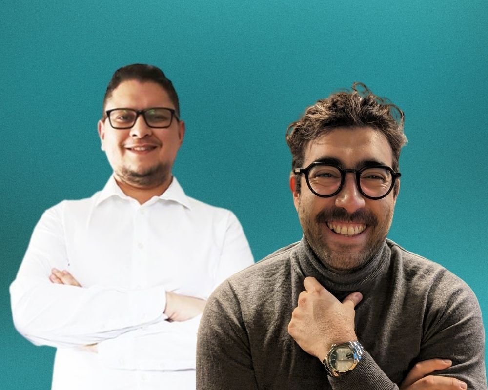 Two smiling men with glasses pose in front of a teal background.
